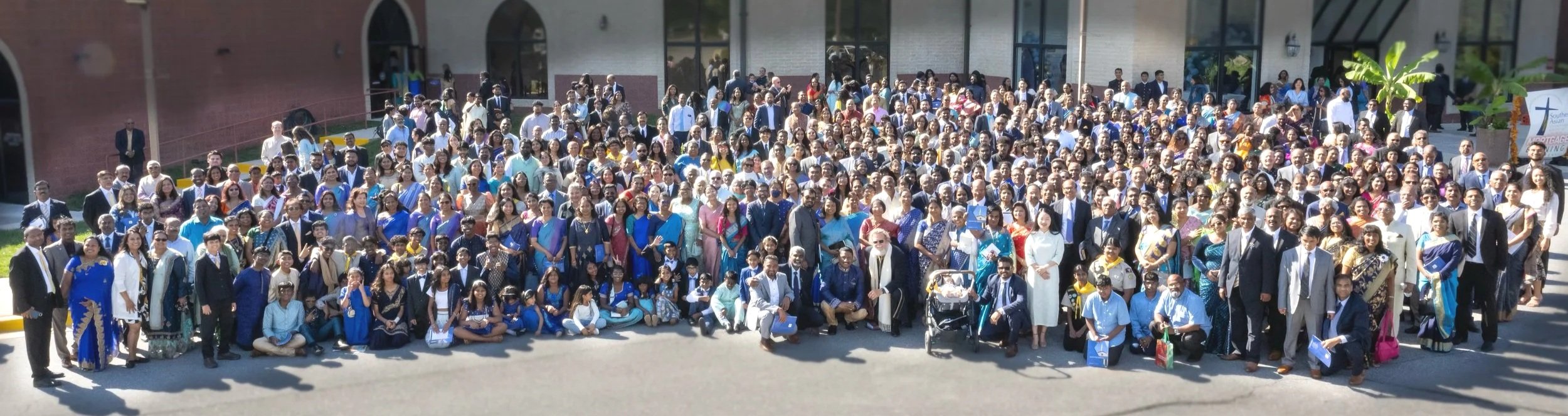 A large group of people gathered outside a building, posing for a photo during a formal event. Many are dressed in traditional and professional attire, with some children in front. The setting appears to be sunny, and there is greenery and a building with large windows in the background.