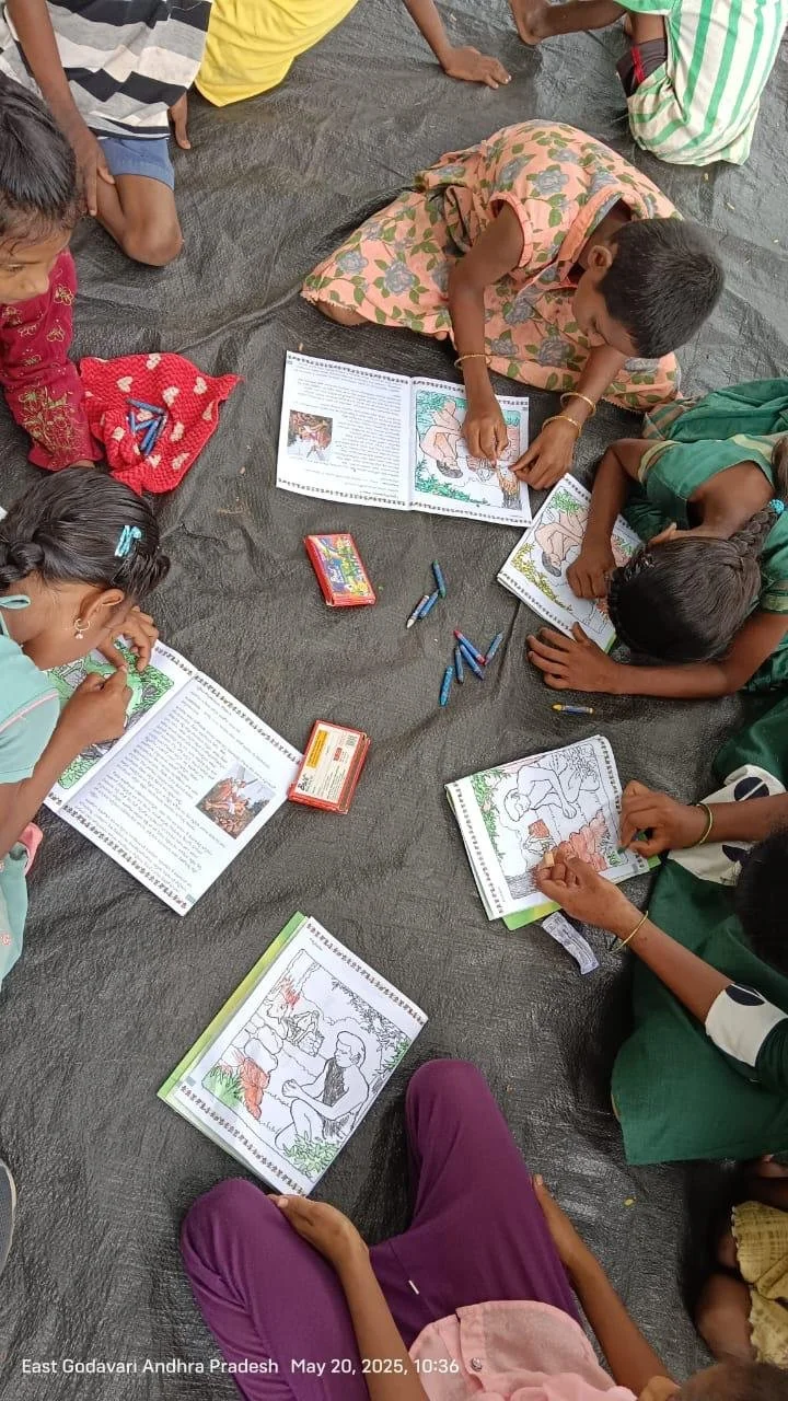 Children sitting on a black mat, coloring and reading illustrated books about traditional stories, outdoors in East Godavari, Andhra Pradesh, on May 20, 2025.