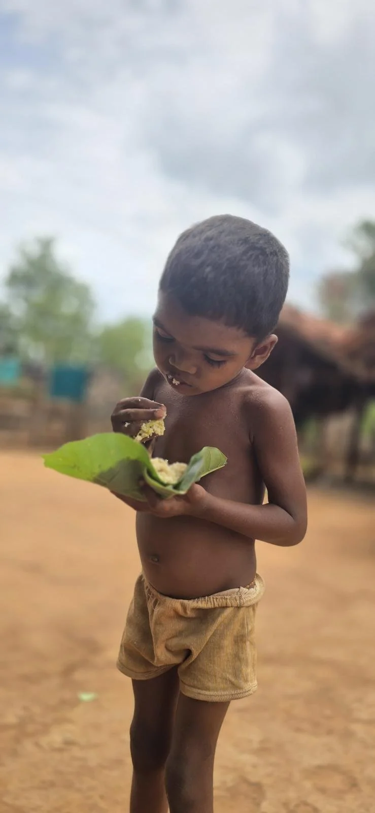 A young boy with short hair and no shirt is holding a leaf with food on it, eating with his right hand, outdoors on a dirt ground with trees and buildings in the background.