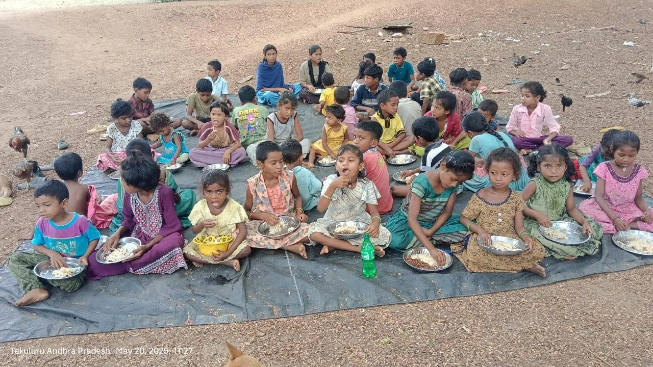 Group of children sitting on black tarpaulin outdoors, eating from metal plates, with some birds nearby in Andhra Pradesh, India.
