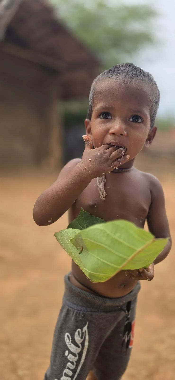 A young boy with short hair is standing outdoors, holding a large green leaf in front of his belly with one hand. He has the other hand near his mouth, with some food or dirt on his fingers and face, and appears to be eating or cleaning his mouth. Th