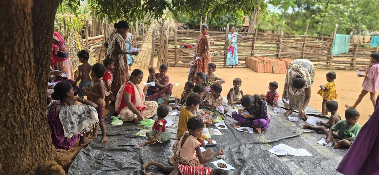 A group of children and women sitting and standing on a black tarp on the ground outdoors, with wooden fences and trees in the background, in a rural setting.