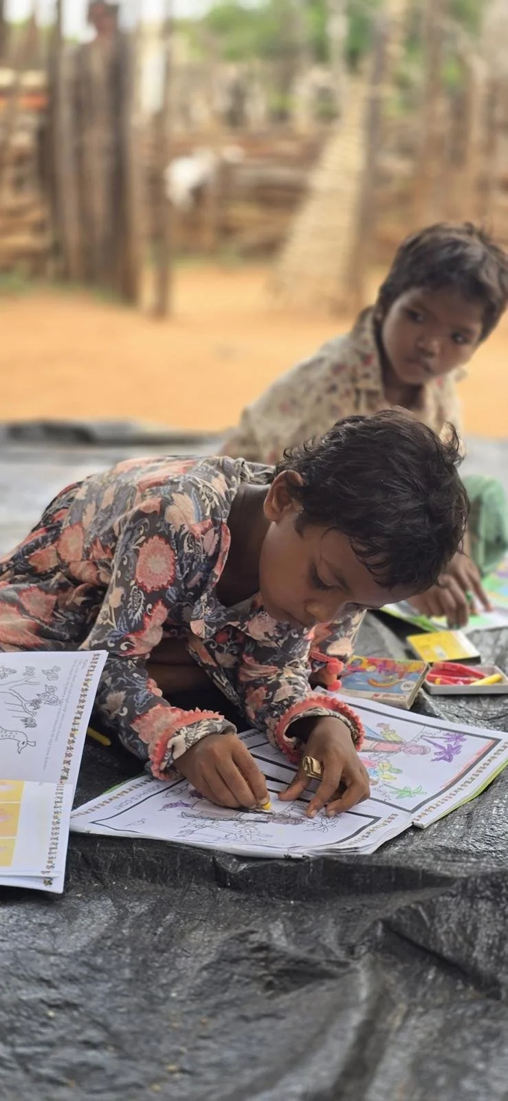 Two young boys sitting on a black plastic sheet outside, coloring and drawing in coloring books. The boy in the foreground is focused on coloring, wearing a floral shirt, and the boy in the background looks on, wearing a patterned shirt. The backgrou