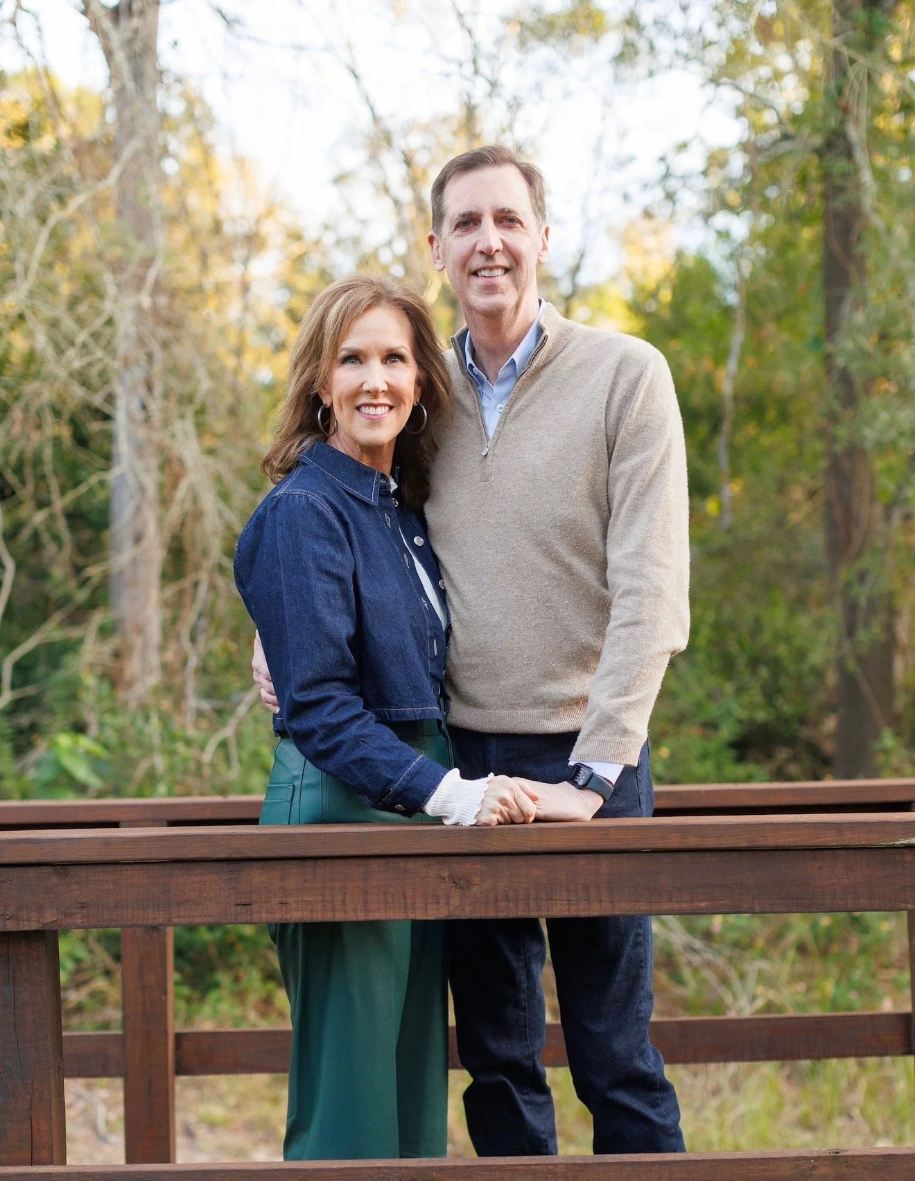 A smiling middle-aged couple standing outdoors behind a wooden railing, surrounded by trees with fall foliage. The woman has shoulder-length brown hair, wears a denim jacket and green pants, and is clasping her hands on the railing. The man has short brown hair, wears a beige sweater over a light blue shirt, and dark pants. They are holding hands and looking at the camera.