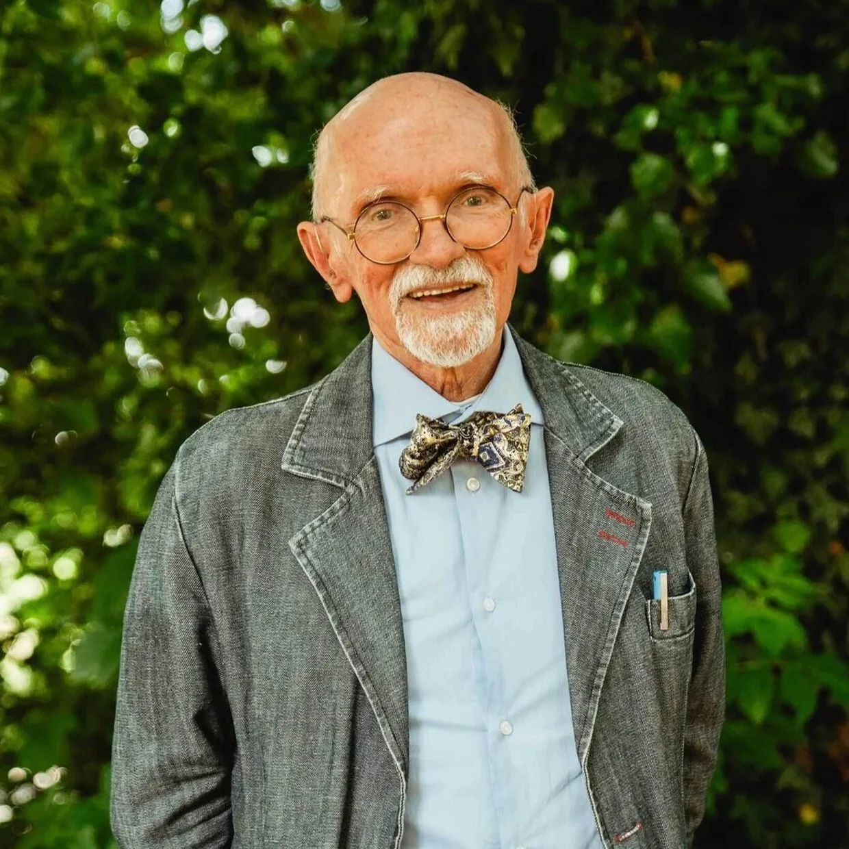 Un uomo anziano con occhiali, barba bianca e vestiti eleganti, sorride davanti a una vegetazione verde.