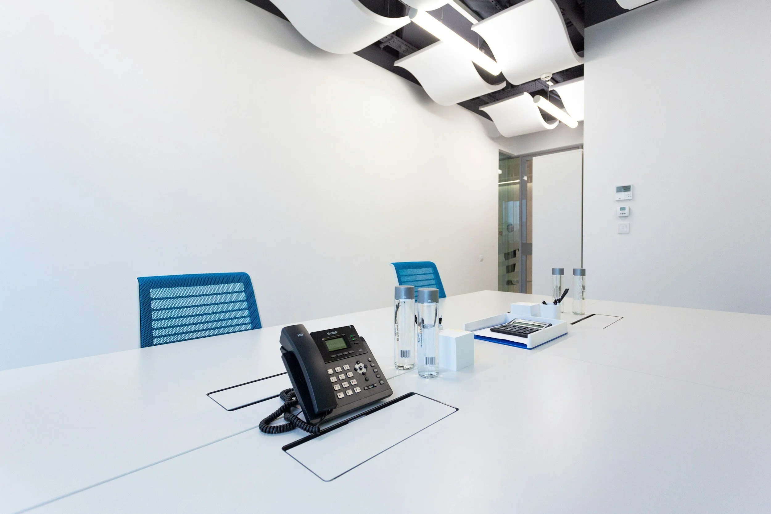 Empty modern conference room with a white table, blue office chairs, a black phone, water bottles, and office supplies.