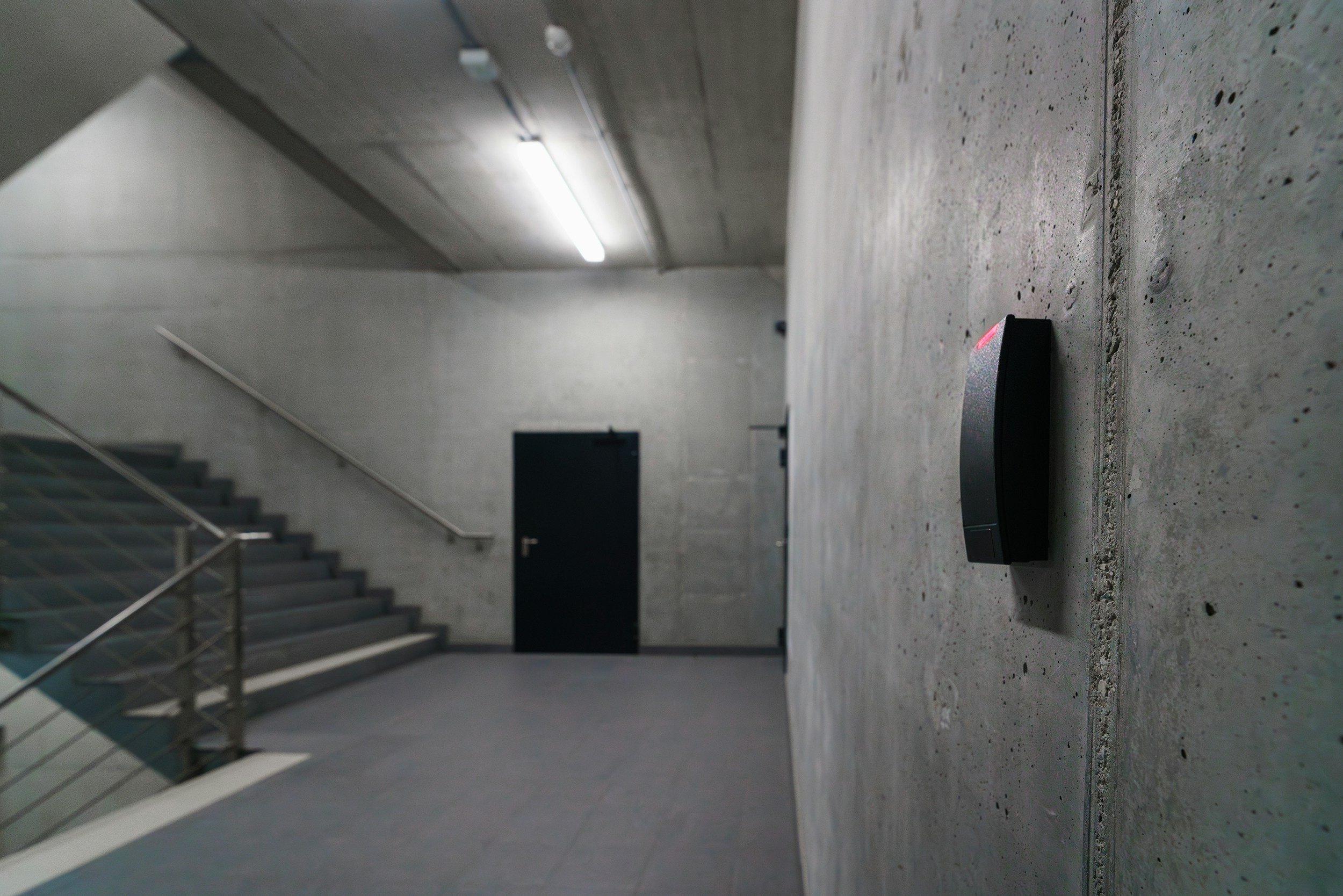 A black automatic hand sanitizer dispenser mounted on a gray concrete wall inside a modern building, with a staircase and a door visible in the background.