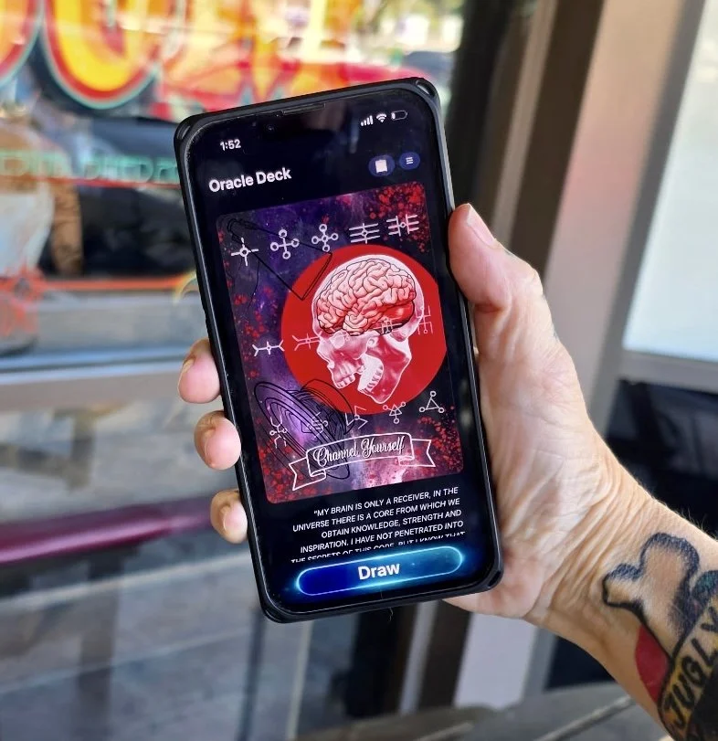 A person holding a smartphone displaying an app called 'The Work Oracle Deck' with a card showing a human brain inside a skull, surrounded by Sacred Circuitry symbols, and the phrase 'Channel Yourself' at the bottom.