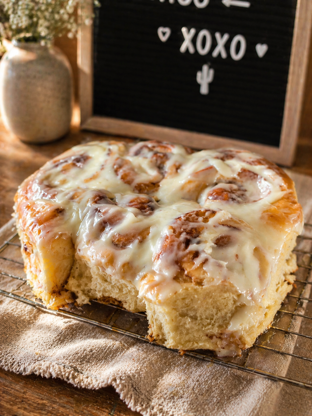 A square pan of freshly baked cinnamon roll cake with cream cheese frosting on a wire rack, with a rustic wooden table, a beige cloth, and a decorative vase with flowers, and a chalkboard sign with 'XOXO' and a cactus in the background.