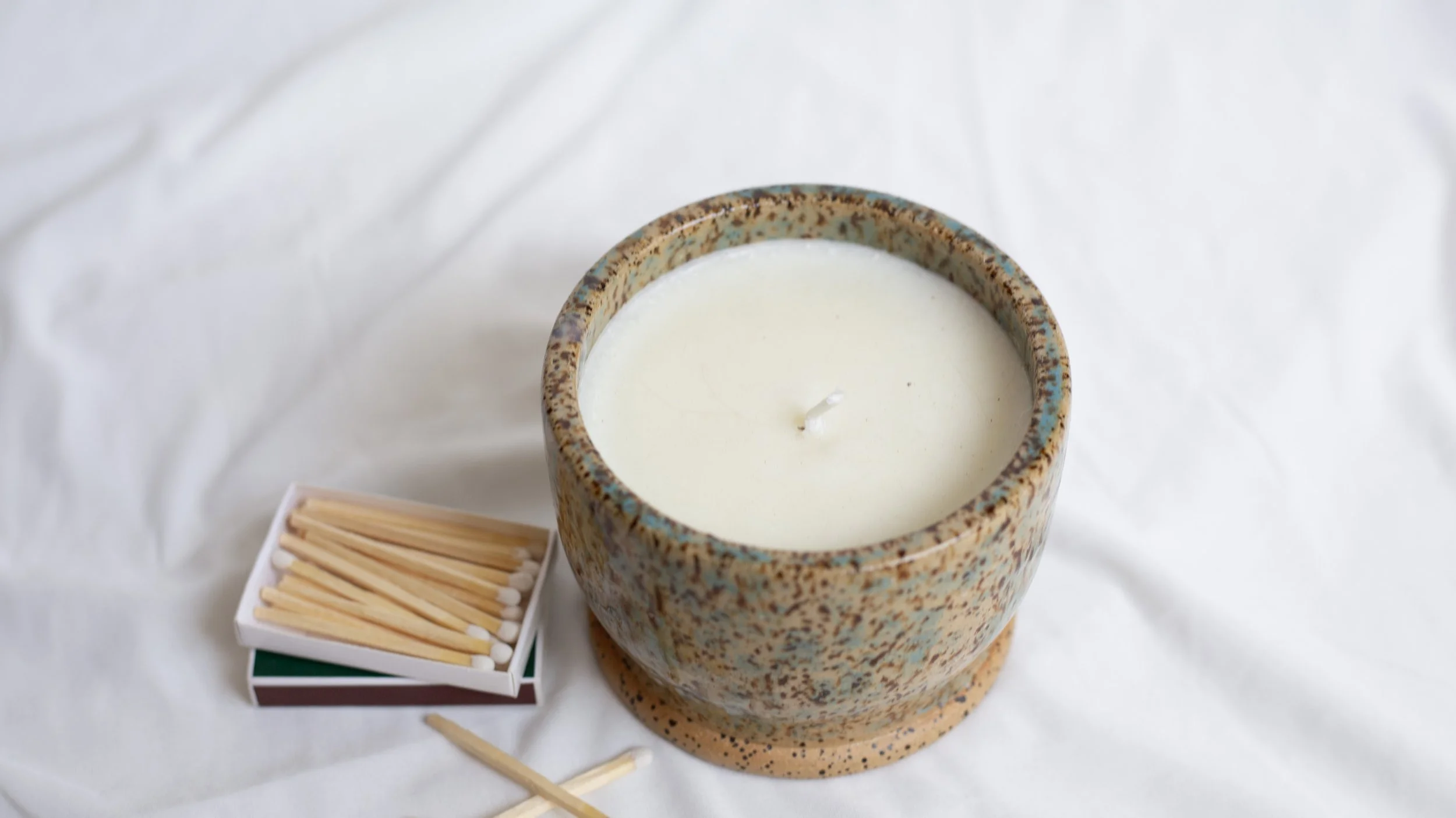 A ceramic candle holder with a white candle inside, placed on a white fabric surface, next to a small box of wooden matches with a matchstick lying nearby.