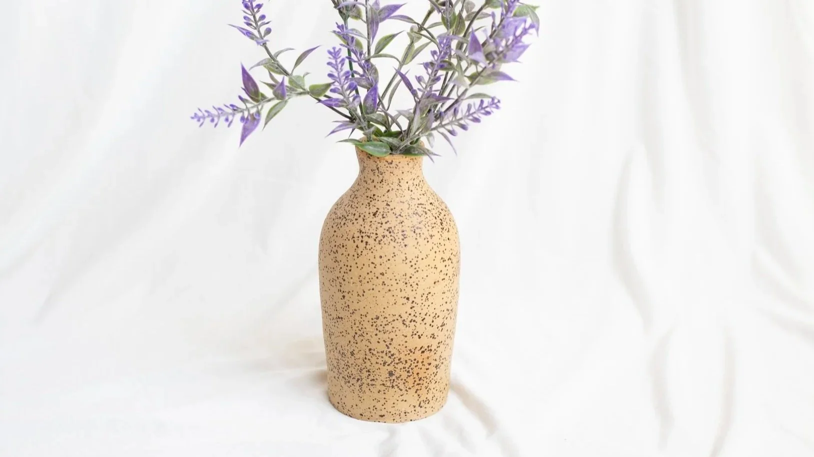 A speckled beige ceramic vase with purple and green foliage. The background is plain white.