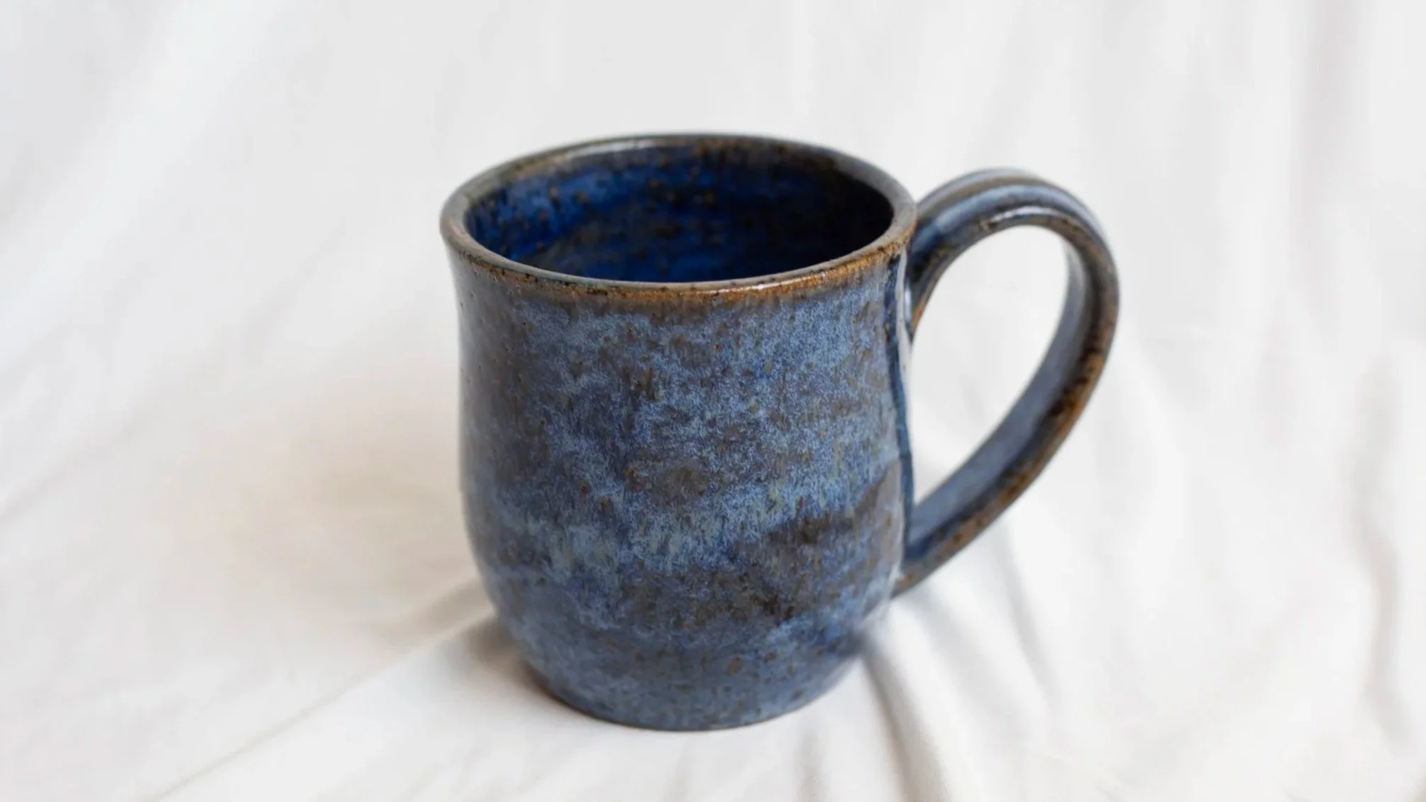 A ceramic mug with a textured, mottled blue glaze on a white fabric background.