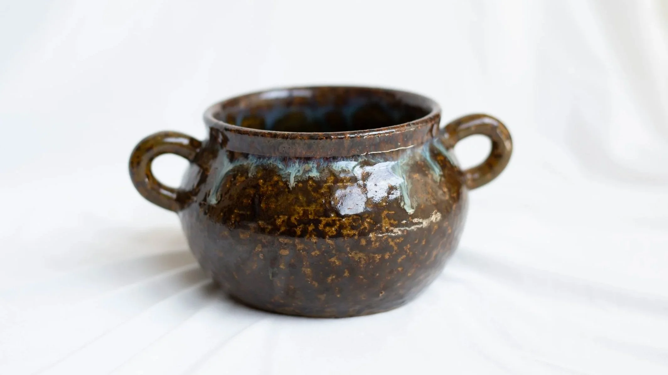 A small ceramic pot with two handles and a glossy dark brown glaze with blue and green streaks, placed on a white background.