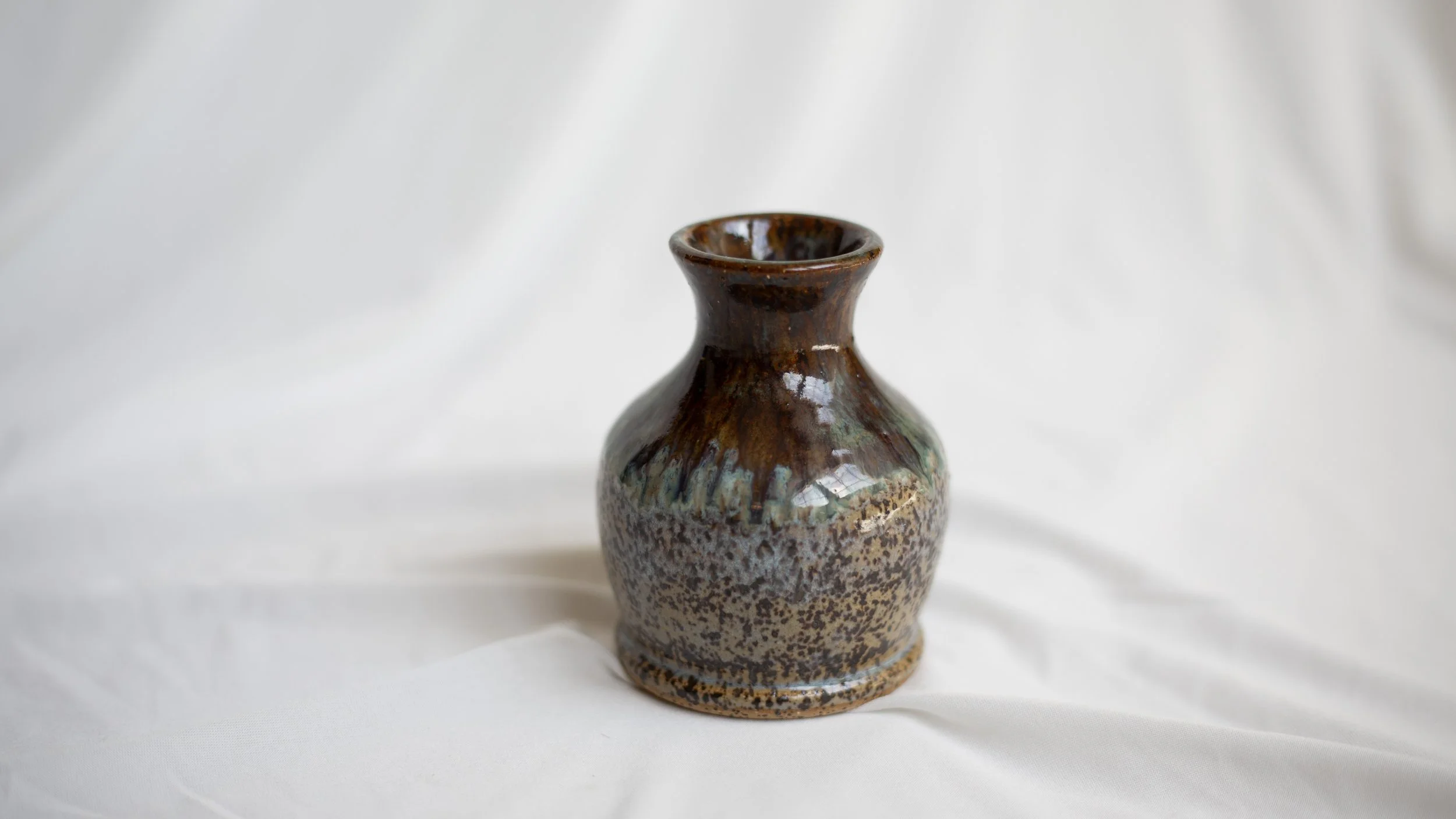 A small ceramic vase with a narrow neck and a rounded body, featuring a multicolored glaze with shades of brown, green, and cream, placed on a white fabric background.