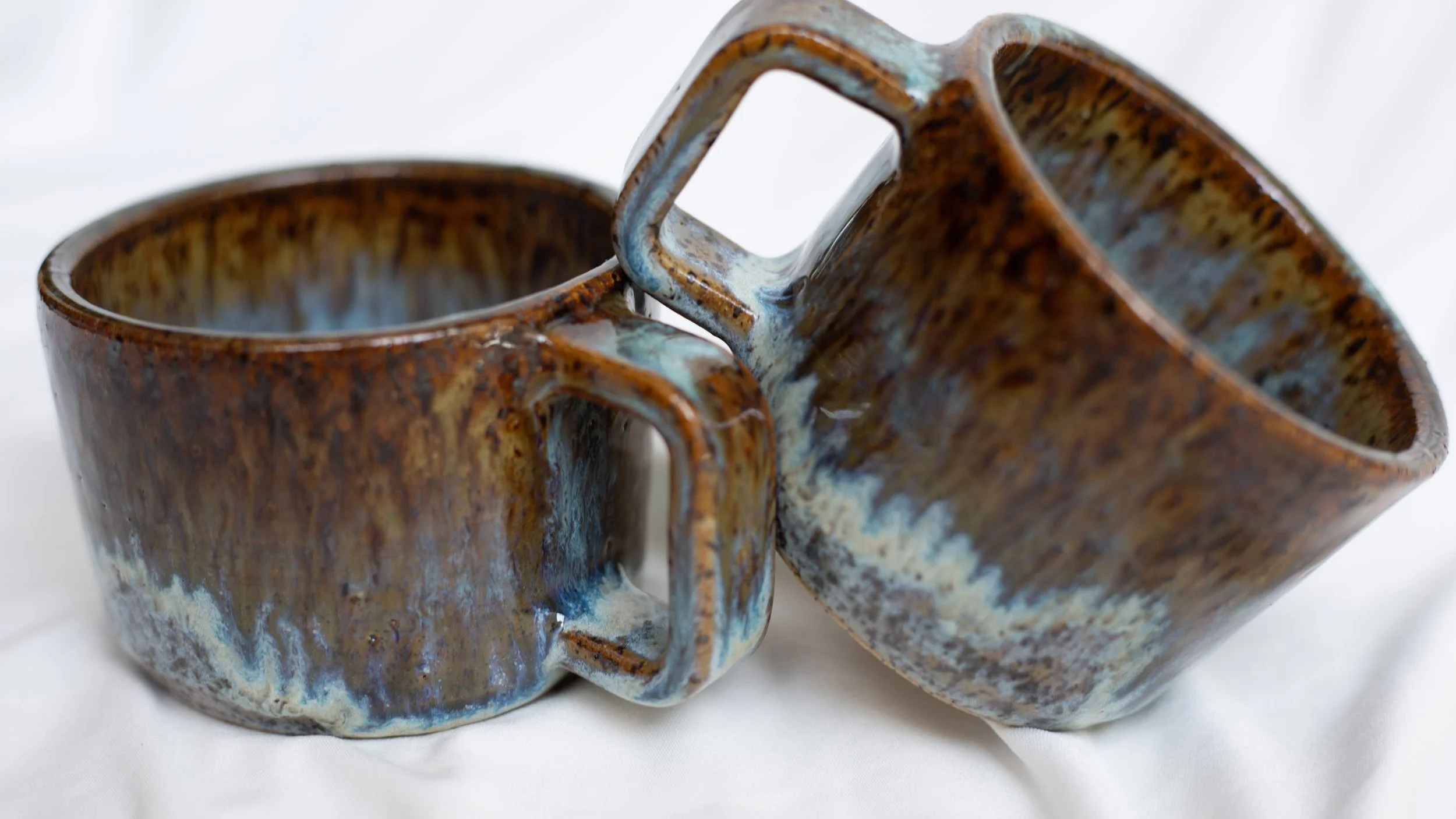 Two handcrafted ceramic mugs, glazed with brown, blue, and cream colors, placed on a white fabric background.