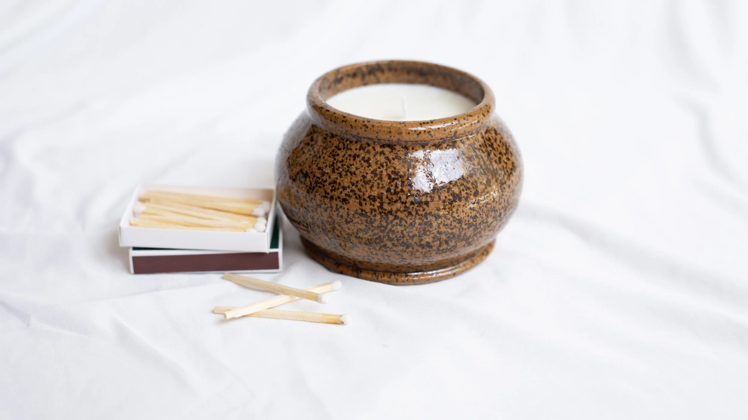A brown ceramic holder with a candle inside, next to a matchbox and some matches on a white cloth surface.