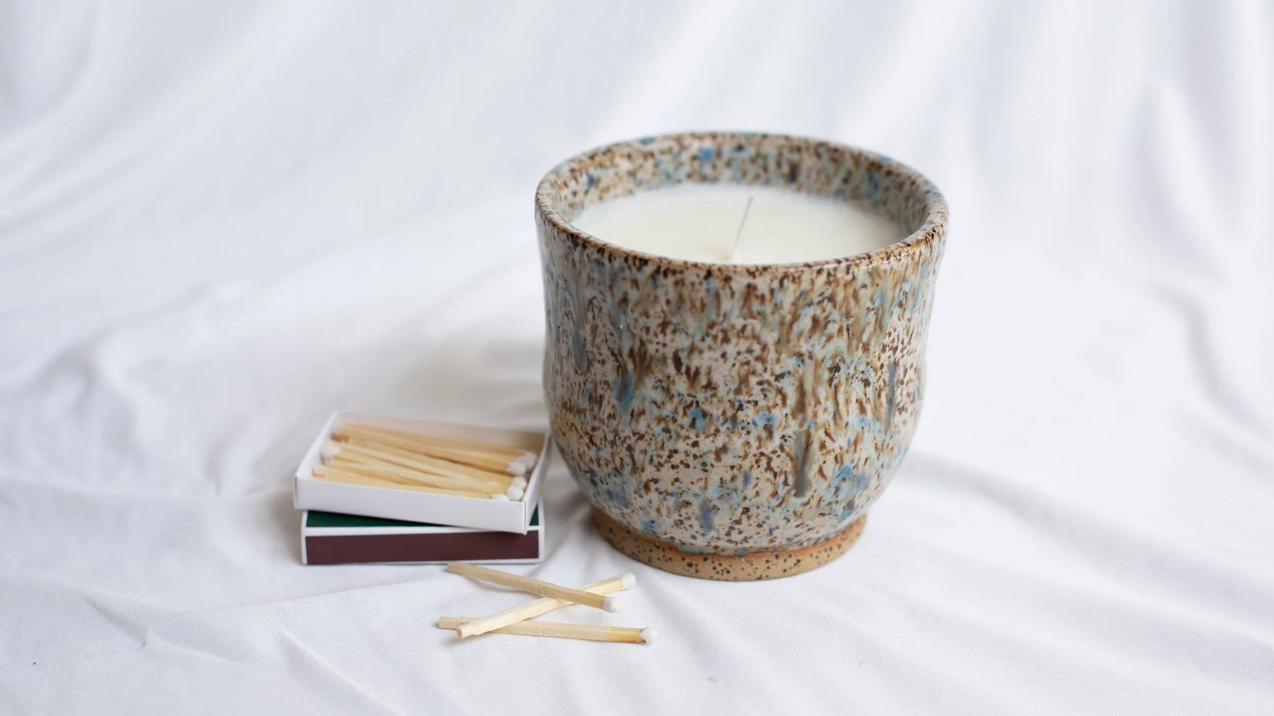 A ceramic candle holder with a white candle inside, beside a small matchbox with matches and a few loose matches on a white fabric background.