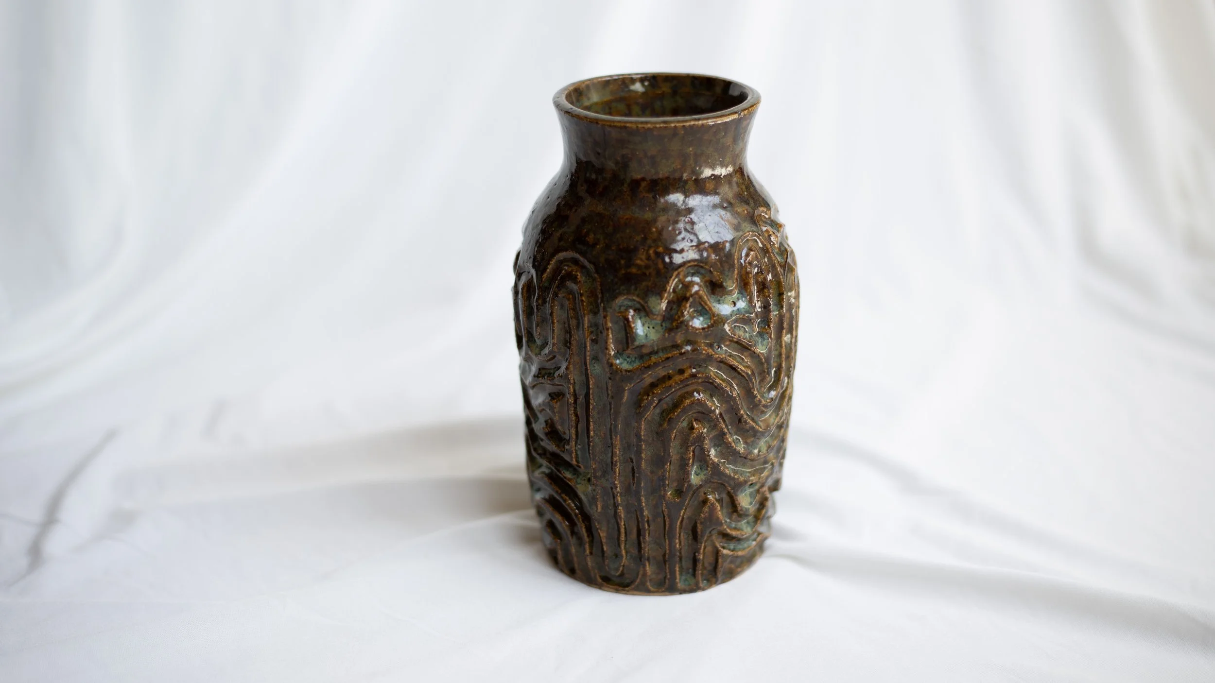 Brown ceramic vase with a textured, abstract design, placed on a white fabric background.