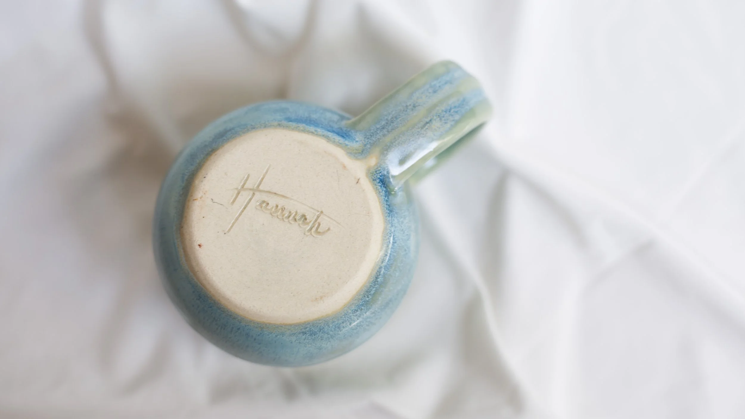 Close-up of a ceramic mug with a glazed blue and green exterior, viewed from the bottom which has the word "Hannah" engraved.