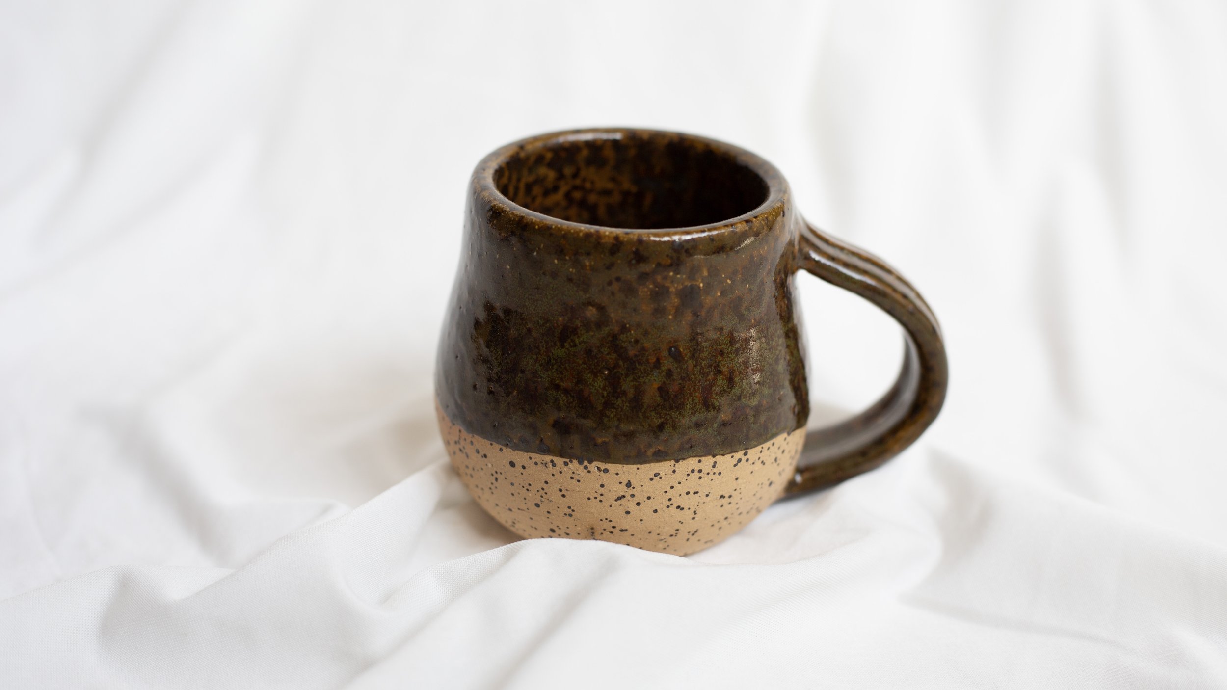 A ceramic mug with a speckled design, featuring a dark brown upper half and a light beige lower half, sitting on a white fabric background.