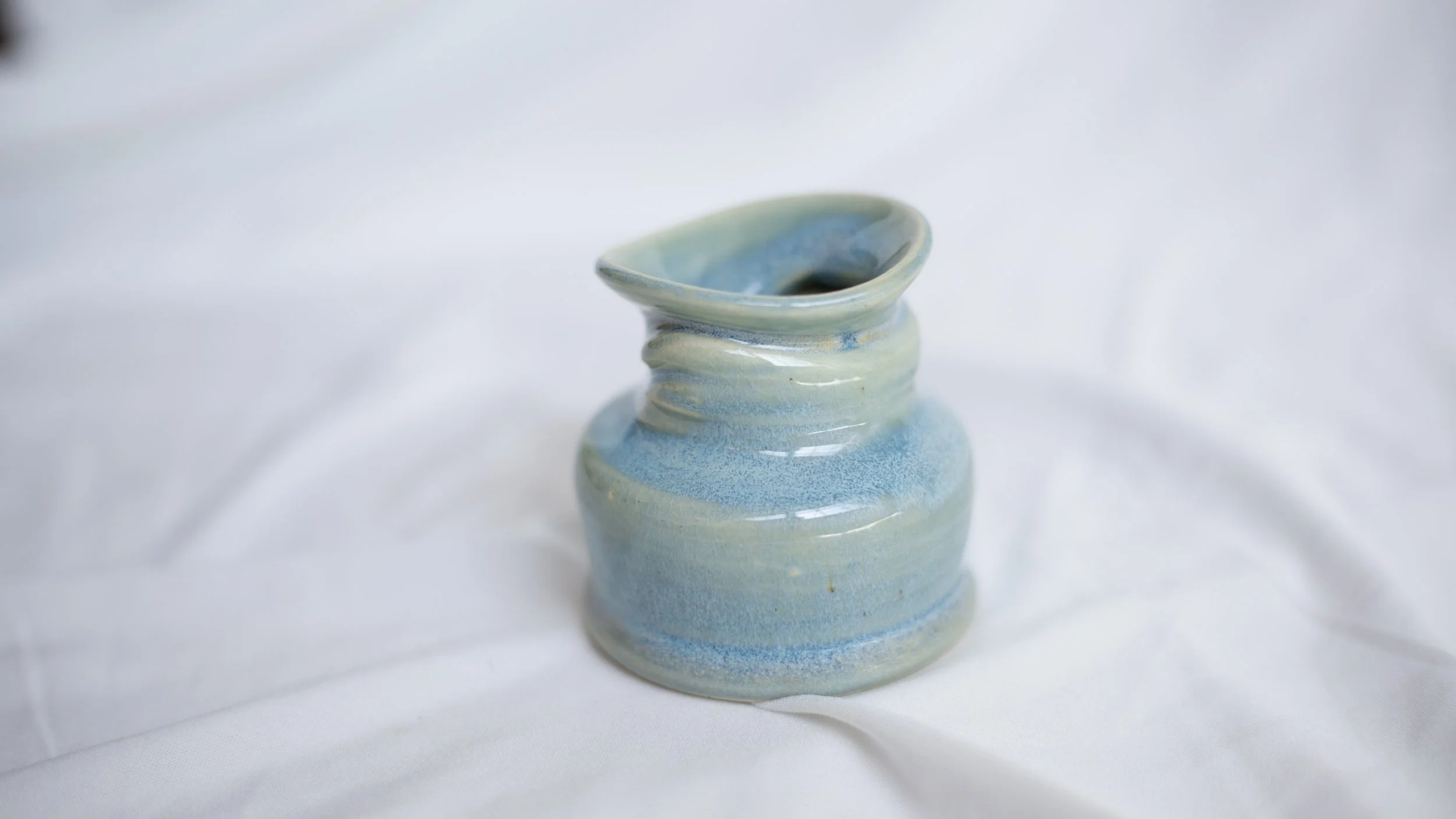 A small ceramic vase with a light blue and green glaze, placed on a white fabric background.