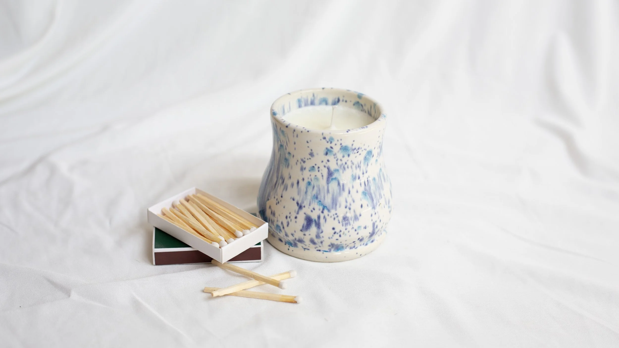 A white and blue speckled candle holder with a lit white candle inside, placed on a white surface, alongside a small deck of matches with some matches spilled out.