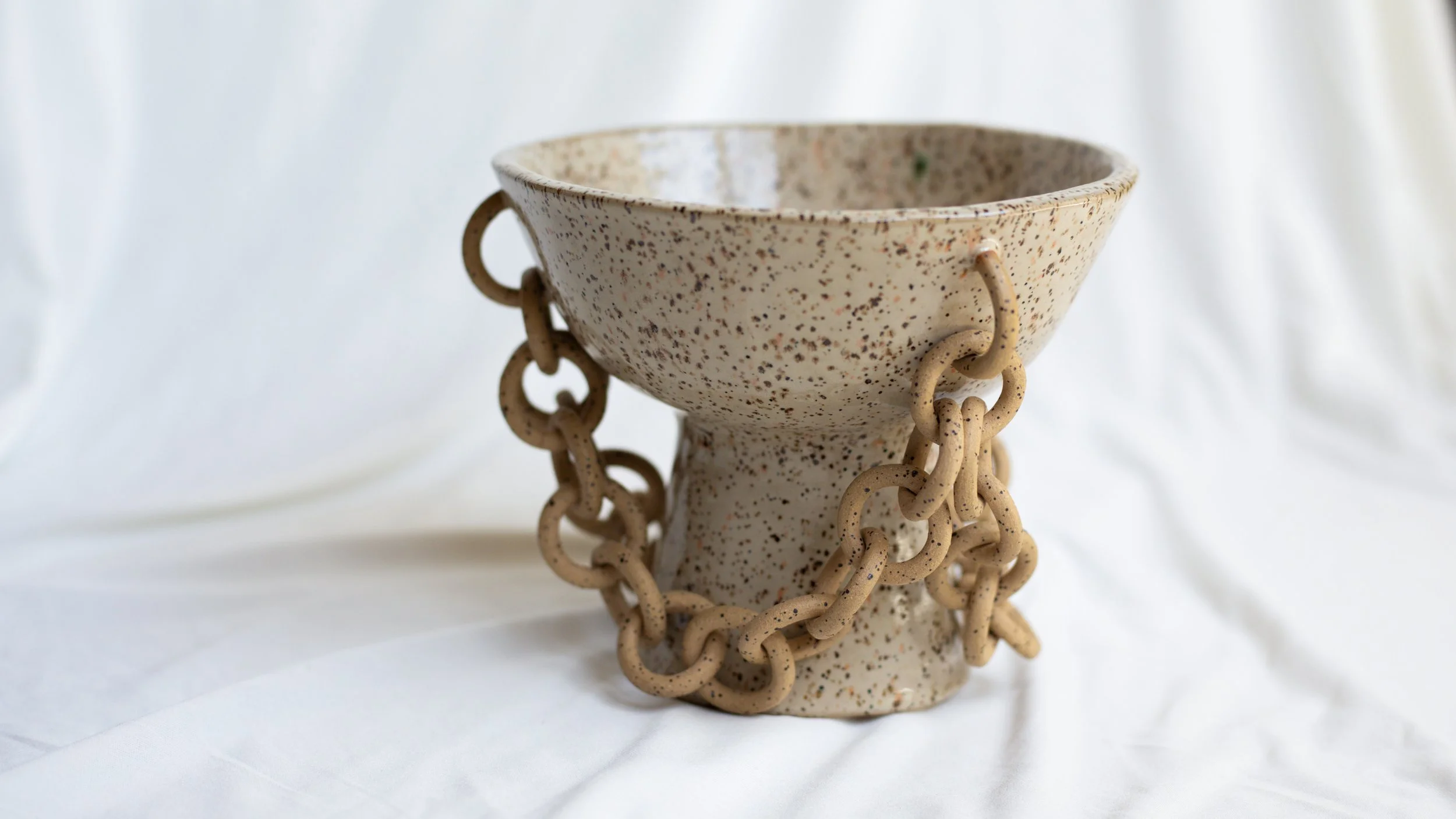 A speckled ceramic bowl with a pedestal base, decorated with a chain of large, beige, textured links hanging around the bowl's rim.