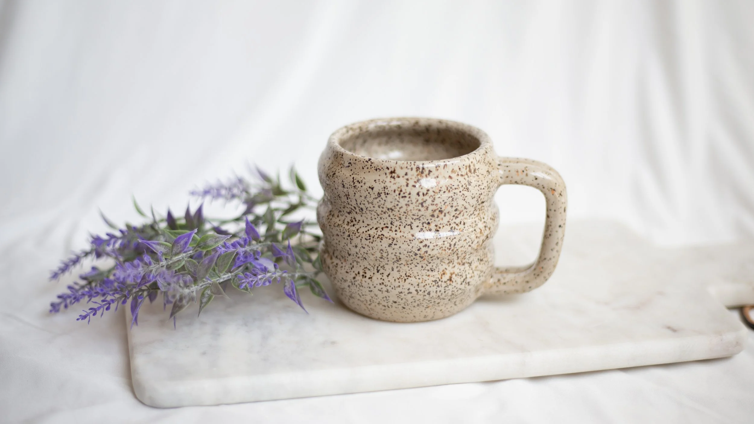 A speckled beige ceramic mug on a white marble tray with purple lavender flowers to the left, set against a white draped background.