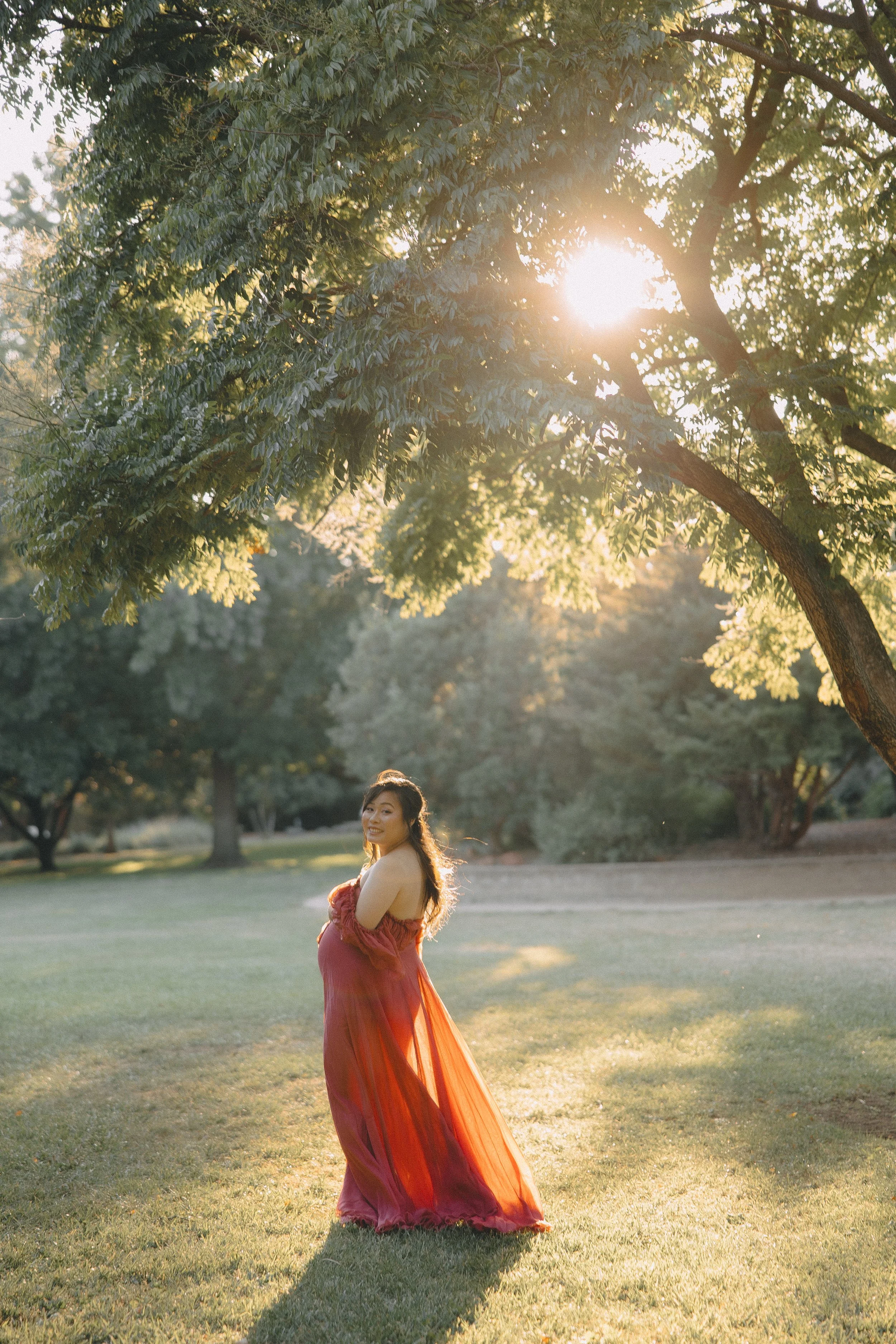 A woman in a flowing, strapless red dress standing outdoors in a grassy park during sunset, with sunlight filtering through trees.