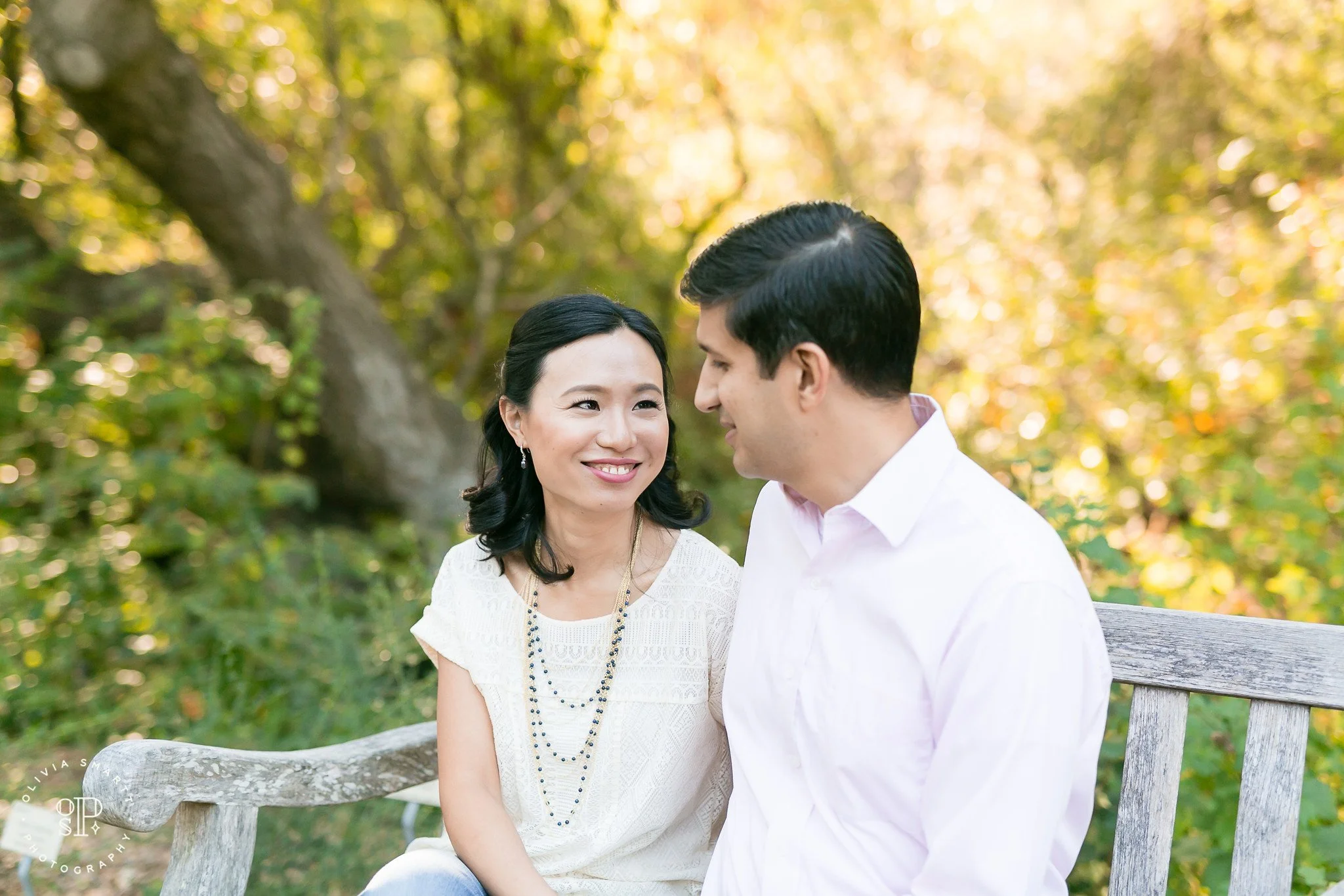 A couple sitting on a wooden bench outdoors, smiling at each other, surrounded by greenery and trees with autumn leaves.