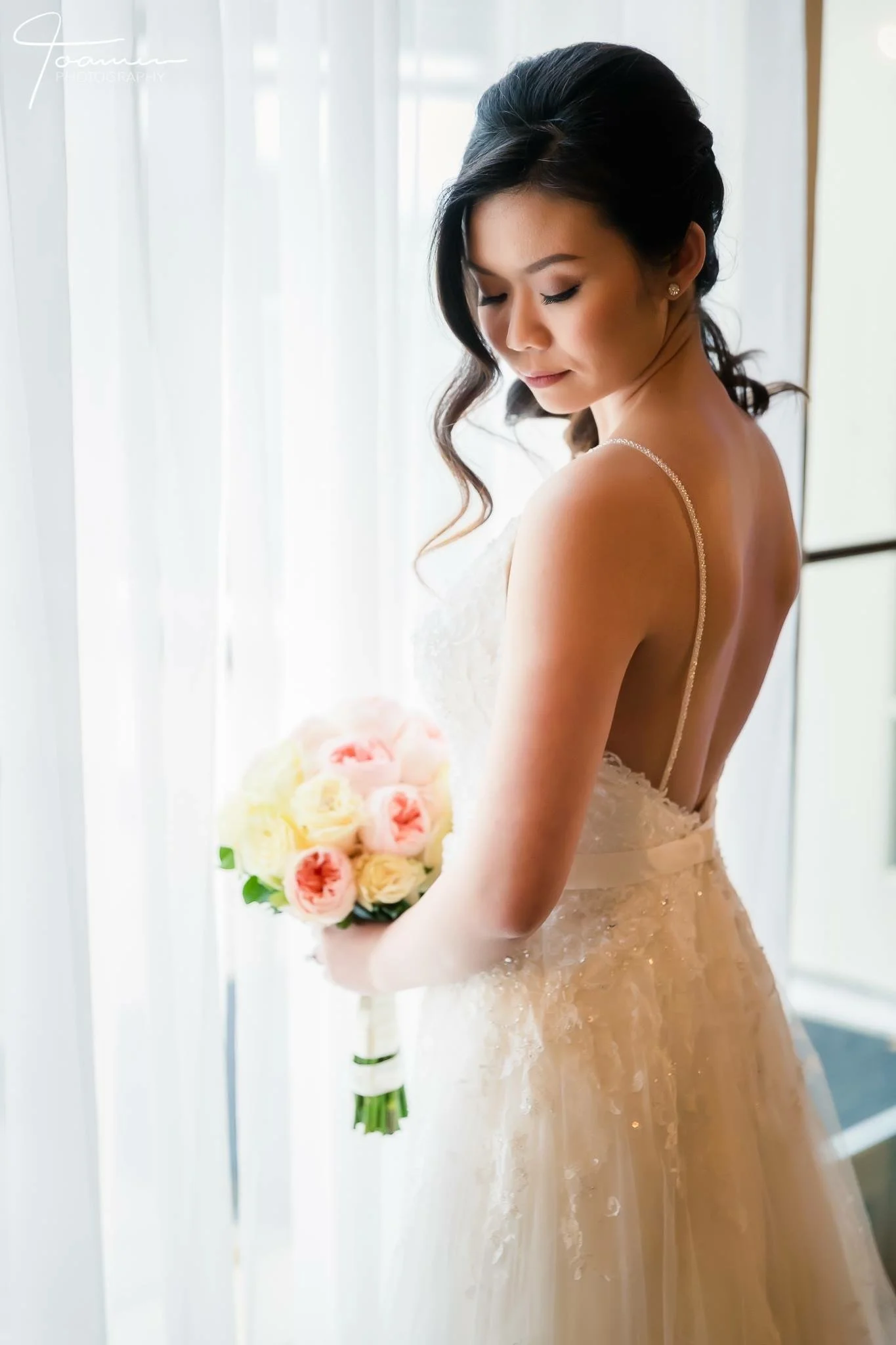 A bride in a white wedding dress holding a bouquet of pink and cream roses, standing near a window with white curtains.
