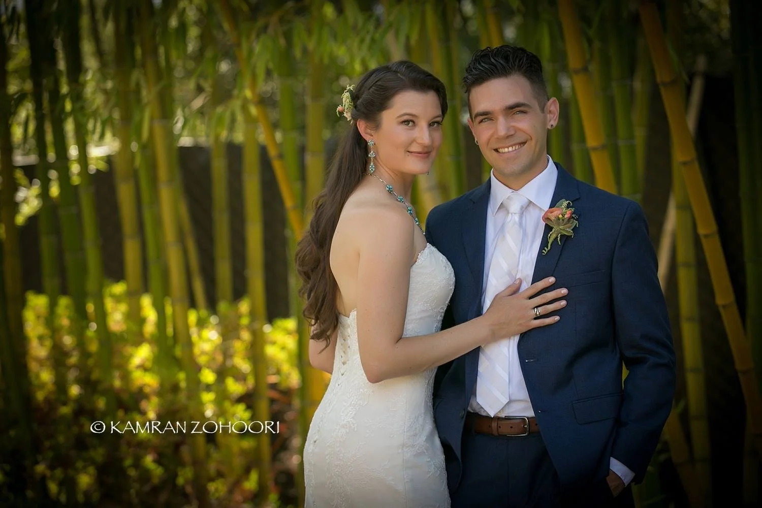 A newlywed couple smiling in a tropical setting with lush green bamboo in the background. The bride is wearing a strapless white lace wedding gown with jewelry, and the groom is in a dark blue suit with a white shirt, tie, and a boutonniere.