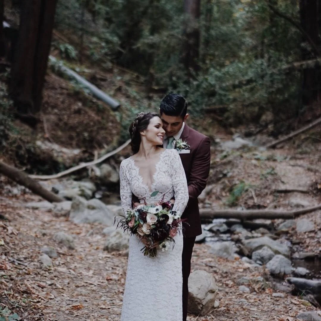 A bride and groom standing close together in a forested area, with the groom embracing the bride from behind. The bride is holding a bouquet of flowers and wearing a white lace wedding dress, while the groom is dressed in a dark suit. They are smilin