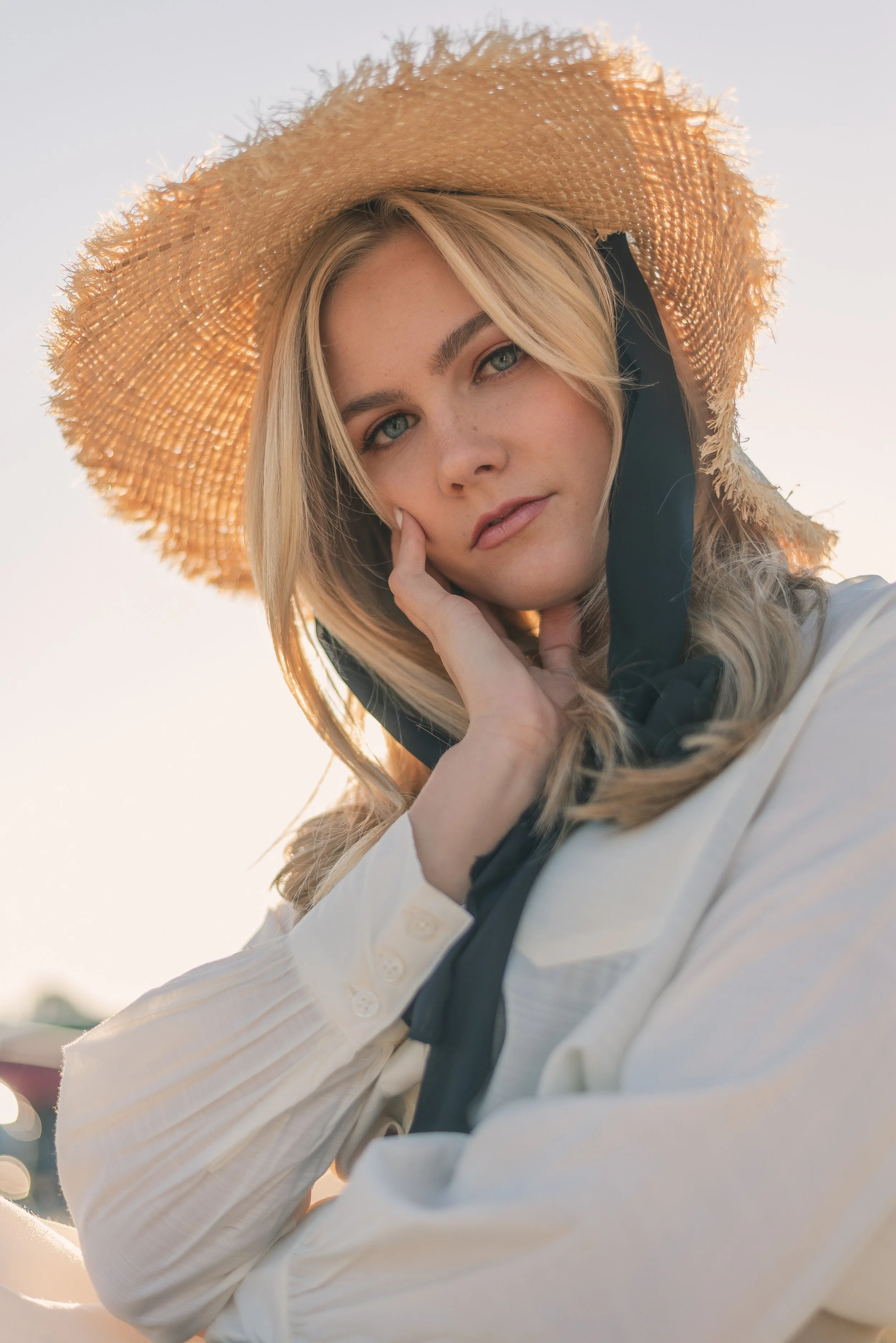 A young woman with blonde hair and blue eyes wearing a wide-brimmed straw hat and a white shirt with black accents, outdoors during sunset.