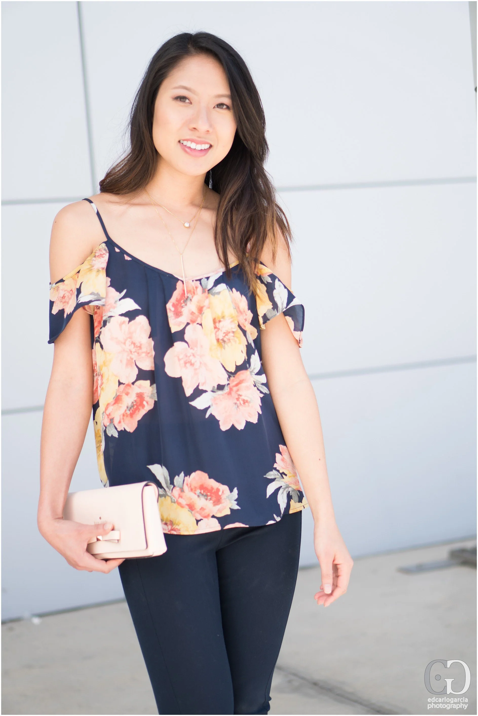 A young woman with long dark hair, smiling, wearing a navy floral off-shoulder top, black pants, holding a beige clutch purse, standing outdoors against a light gray wall.