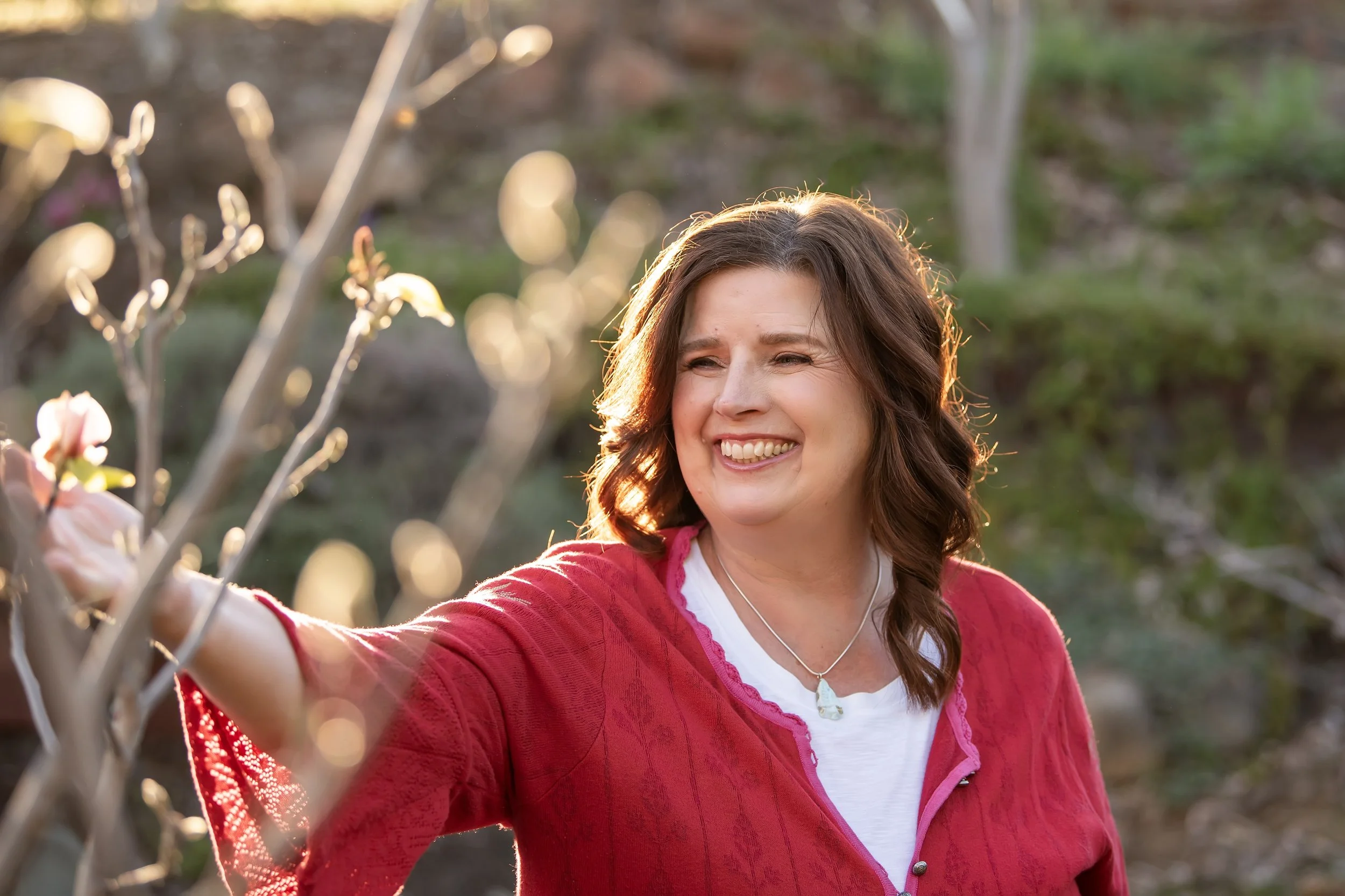 A woman with shoulder-length brown hair smiling outdoors, wearing a red jacket over a white shirt, with a blurred background of trees and sunlight.
