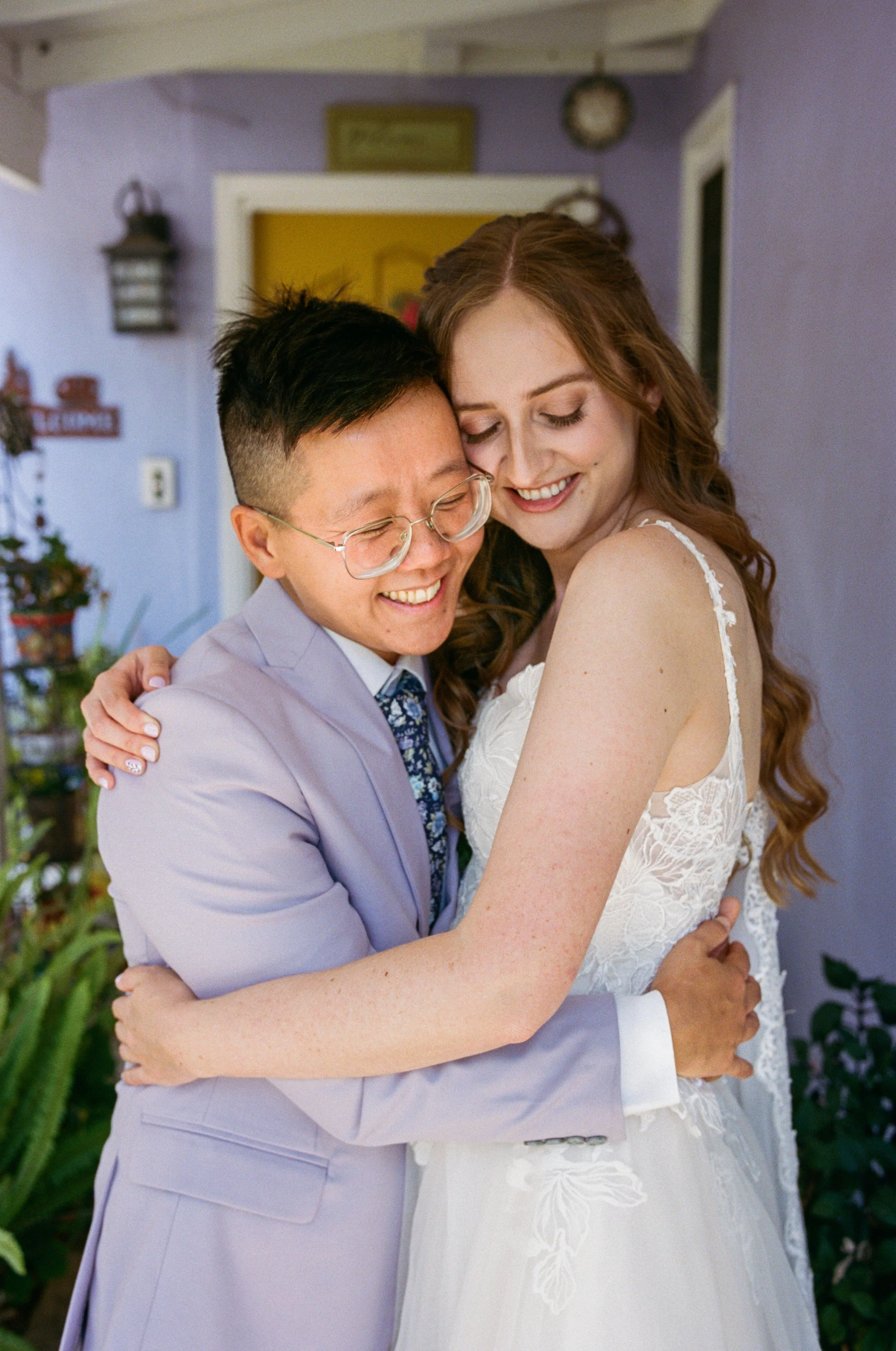 Bride in a suite embracing her bride in a white wedding dress, both smiling with eyes closed, indoors with purple walls and green plants in the background.