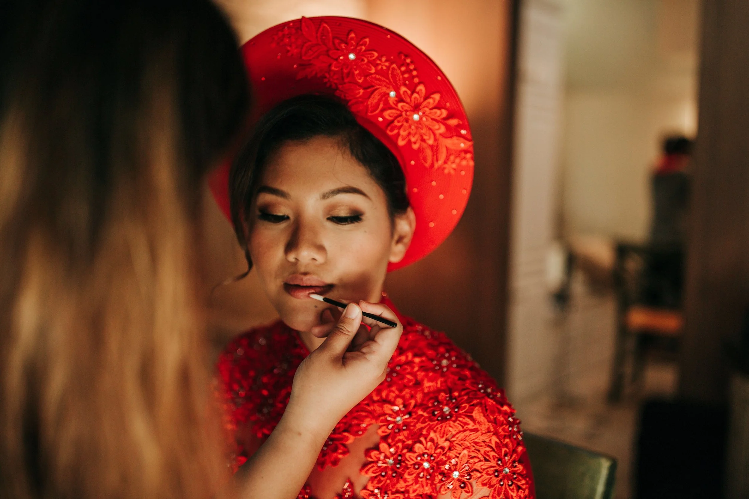A woman in traditional red attire and a large matching hat is having her makeup applied by a makeup artist.