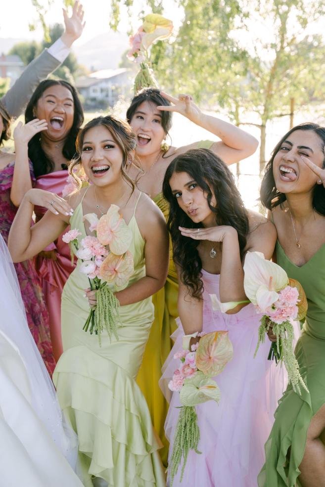 Group of women smiling and posing outdoors at a celebration, wearing colorful dresses, holding bouquets, with trees and sunlight in background.