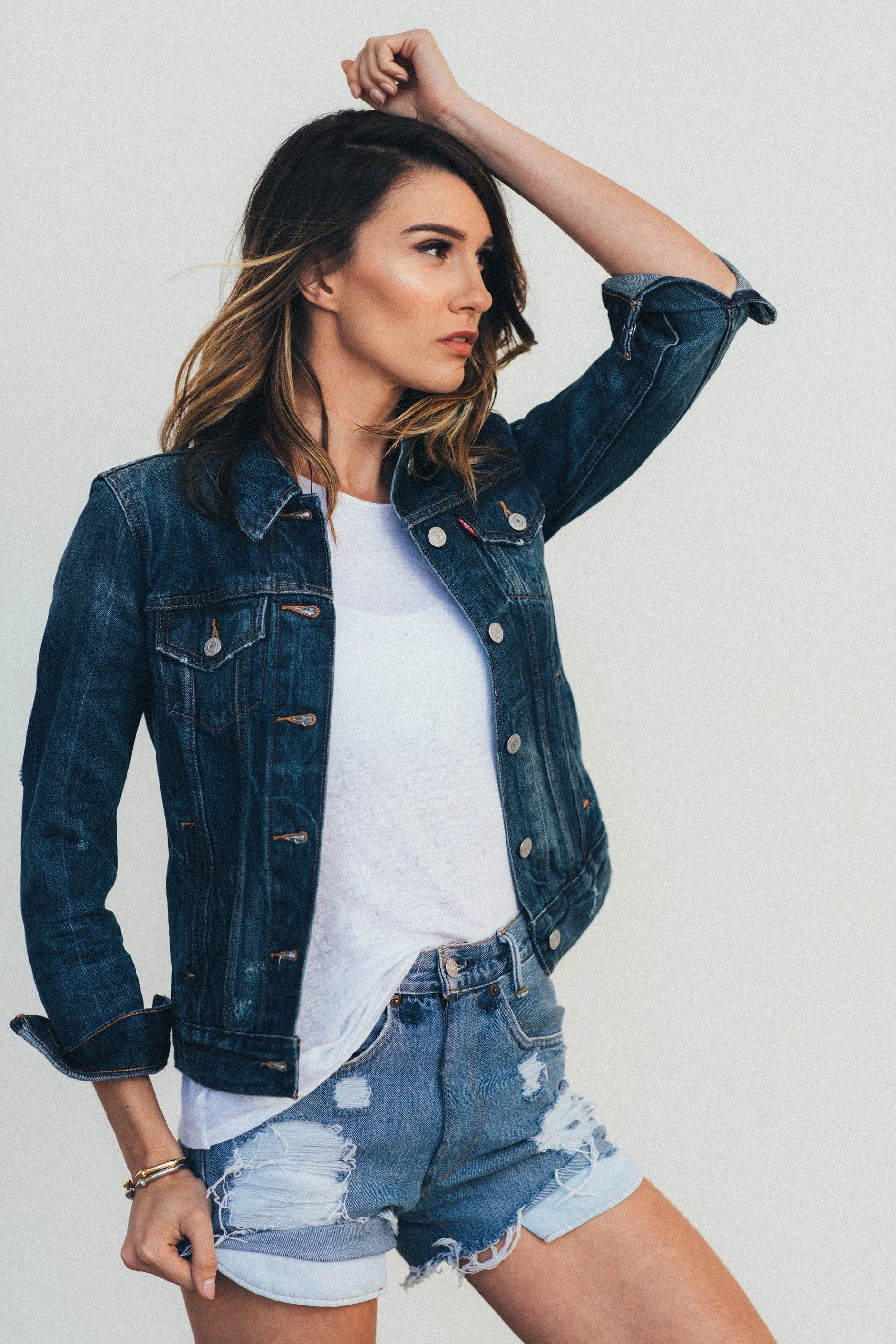 A woman with shoulder-length brown hair wearing a denim jacket, white t-shirt, and distressed denim shorts posing with her right arm raised and left hand holding her shorts.