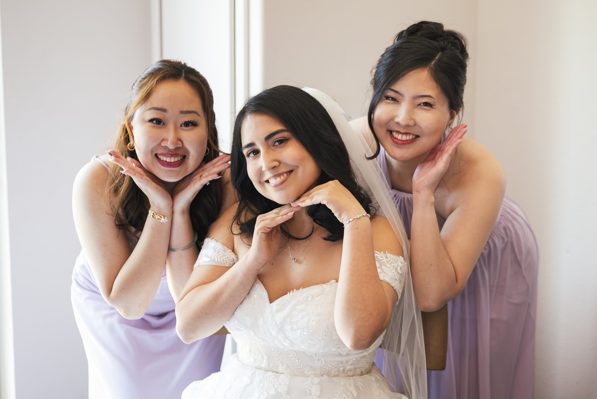 Three women at a wedding, one in a white wedding dress and veil, flanked by two women in lavender dresses, all smiling and looking at the camera with playful poses.