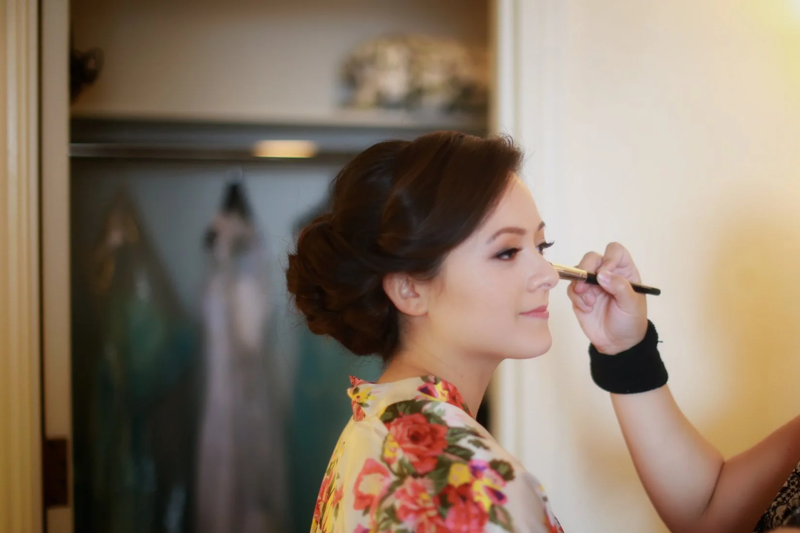 Woman with styled hair getting her makeup done, wearing a floral robe, in a room with a closet in the background.