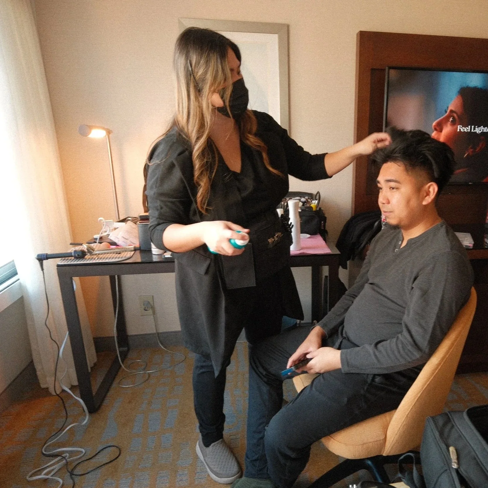 A woman with long hair wearing a black mask is styling a man's hair in a hotel room. The man is seated in a beige chair, holding a phone, and wearing a gray long-sleeve shirt. Behind them is a TV displaying a woman with dark hair, and a table with va