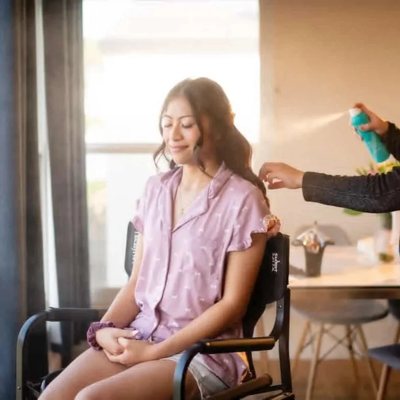 A woman in a pink satin pajama top sitting in a wheelchair, while another woman sprays her hair with a blue spray bottle in a well-lit room.