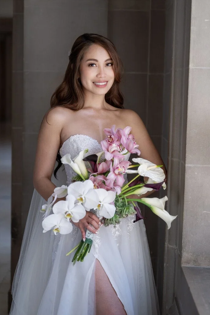 A bride in a strapless white wedding dress with a slit, smiling and holding a large bouquet of pink, white, and dark purple flowers, standing indoors near a wall.