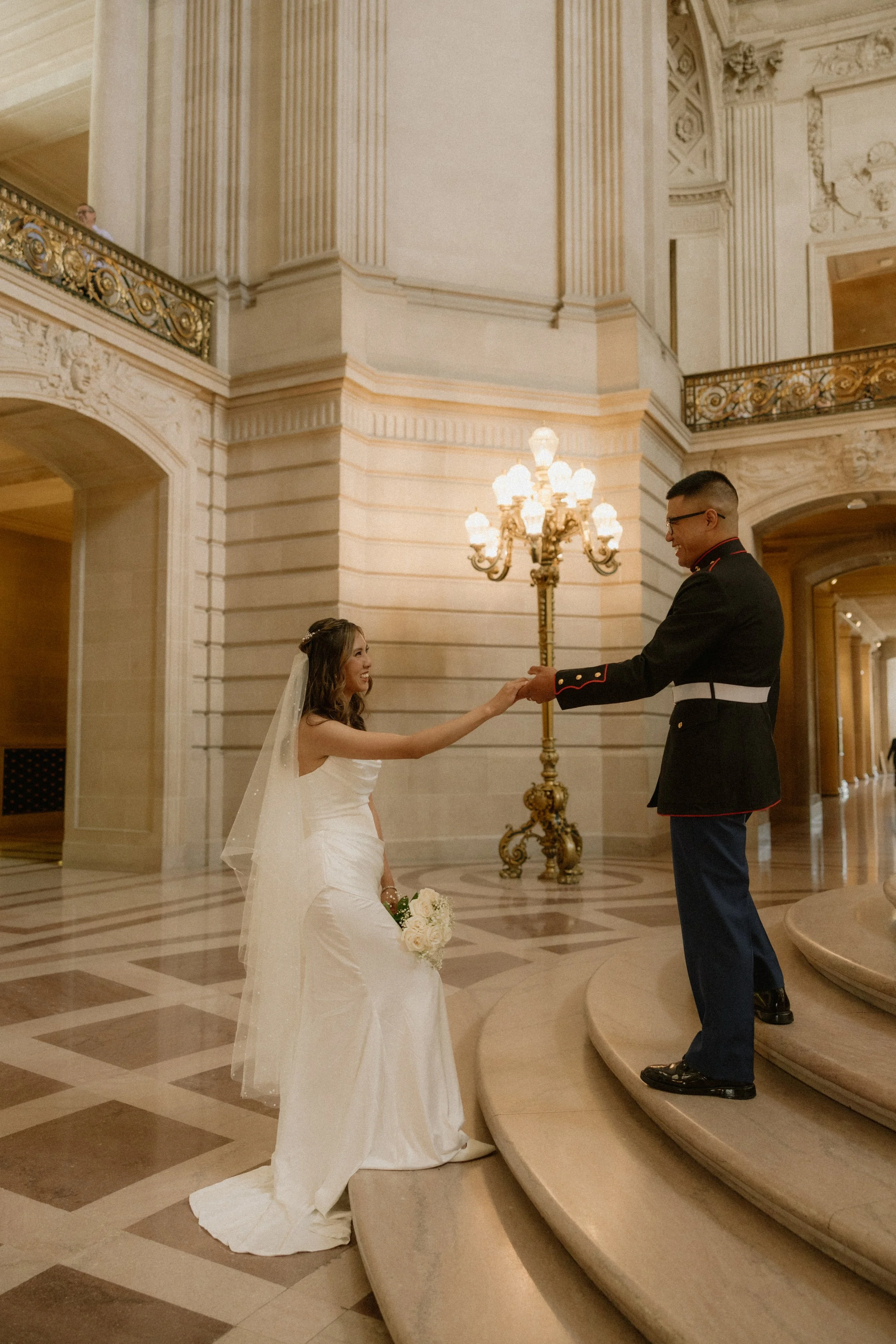 A bride in a white wedding dress with a veil and bouquet smiling at a groom in a formal military uniform on a staircase inside an ornate building.