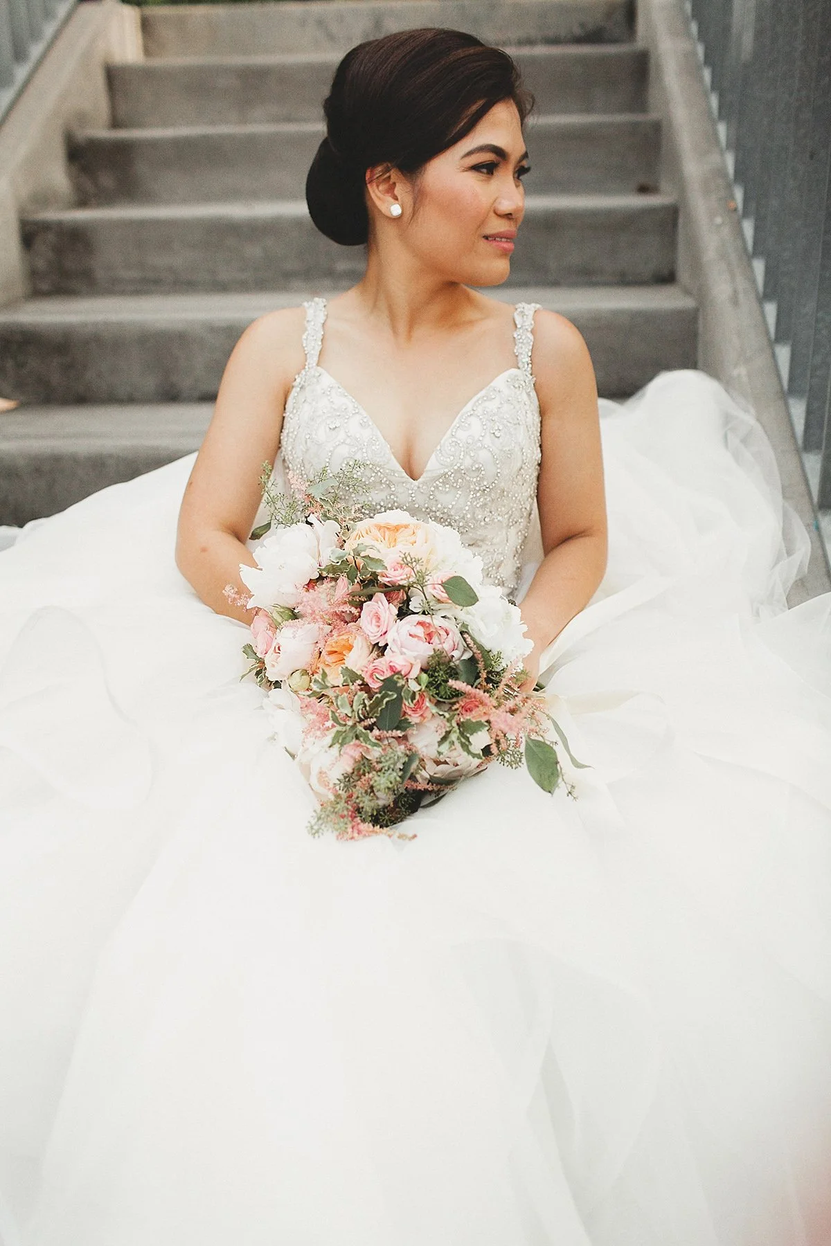 A bride sitting on stairs, holding a pink and white bouquet, wearing a white wedding gown with beadwork, and pearl earrings.
