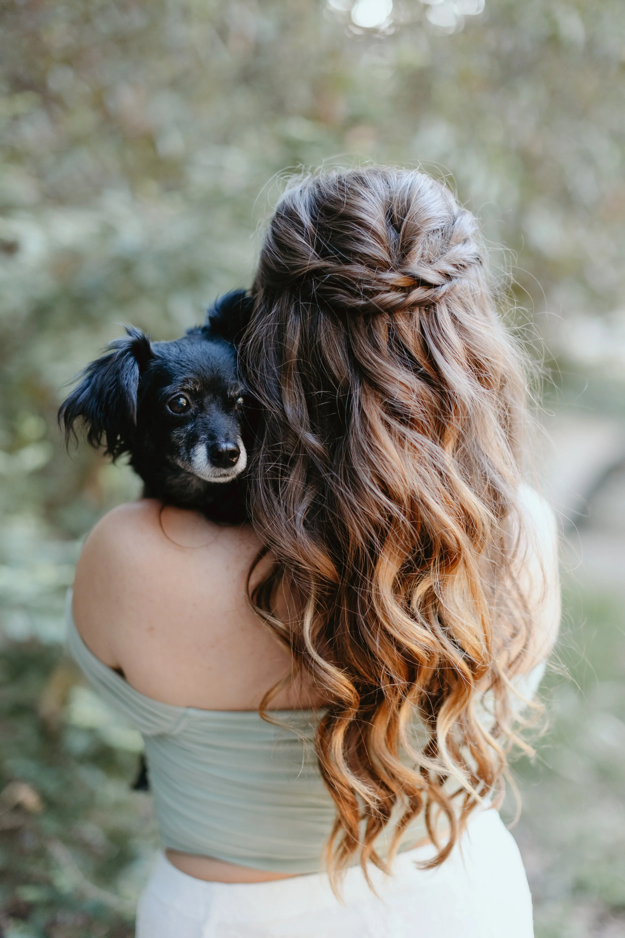 A woman with long, curly, reddish-brown hair styled in a braid, holding a small black and white dog, outdoors with blurred green trees in the background.