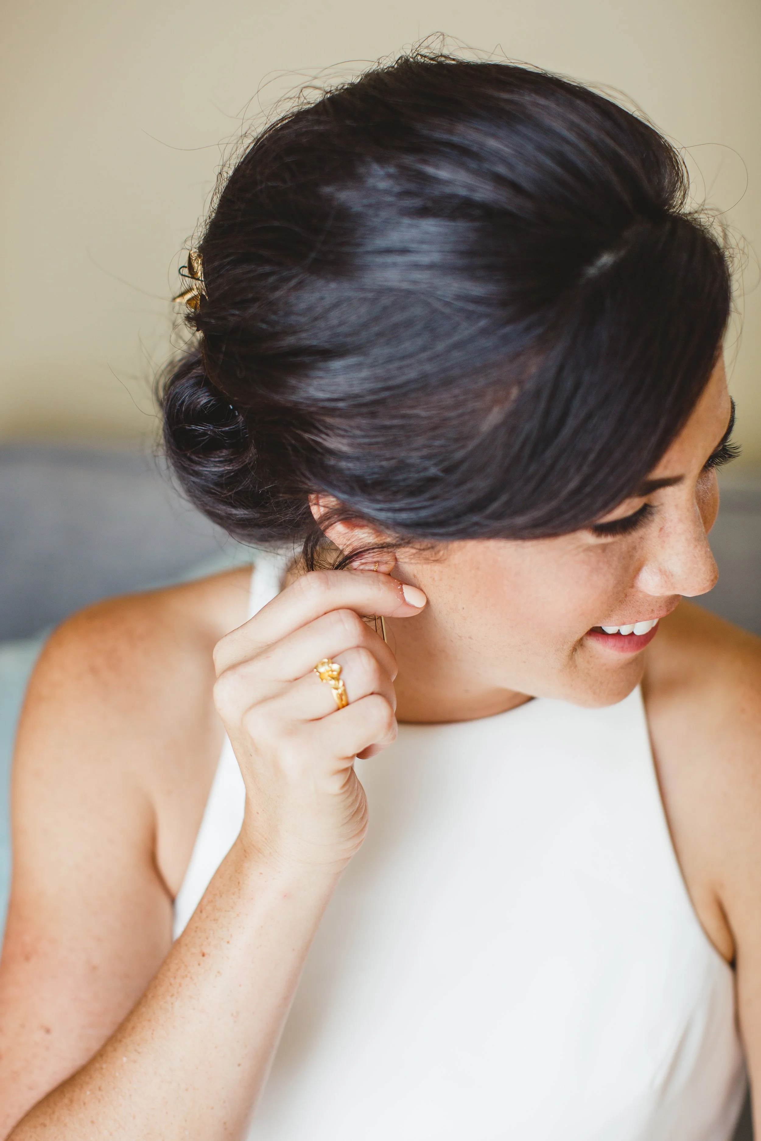 Close-up of a woman with dark hair styled in an elegant bun, wearing a sleeveless white top, adjusting an earring, smiling gently.