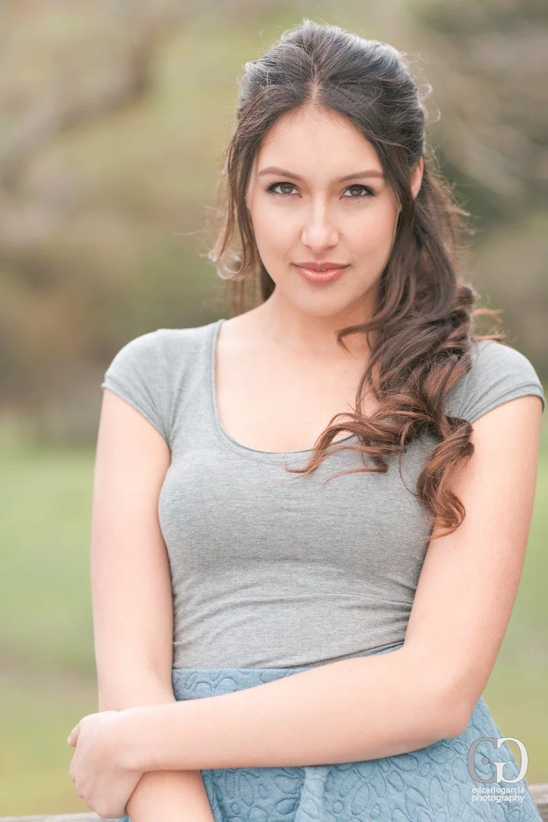 A young woman with long, wavy brown hair tied to one side, wearing a gray t-shirt and a blue skirt, standing outdoors with a blurred natural background.